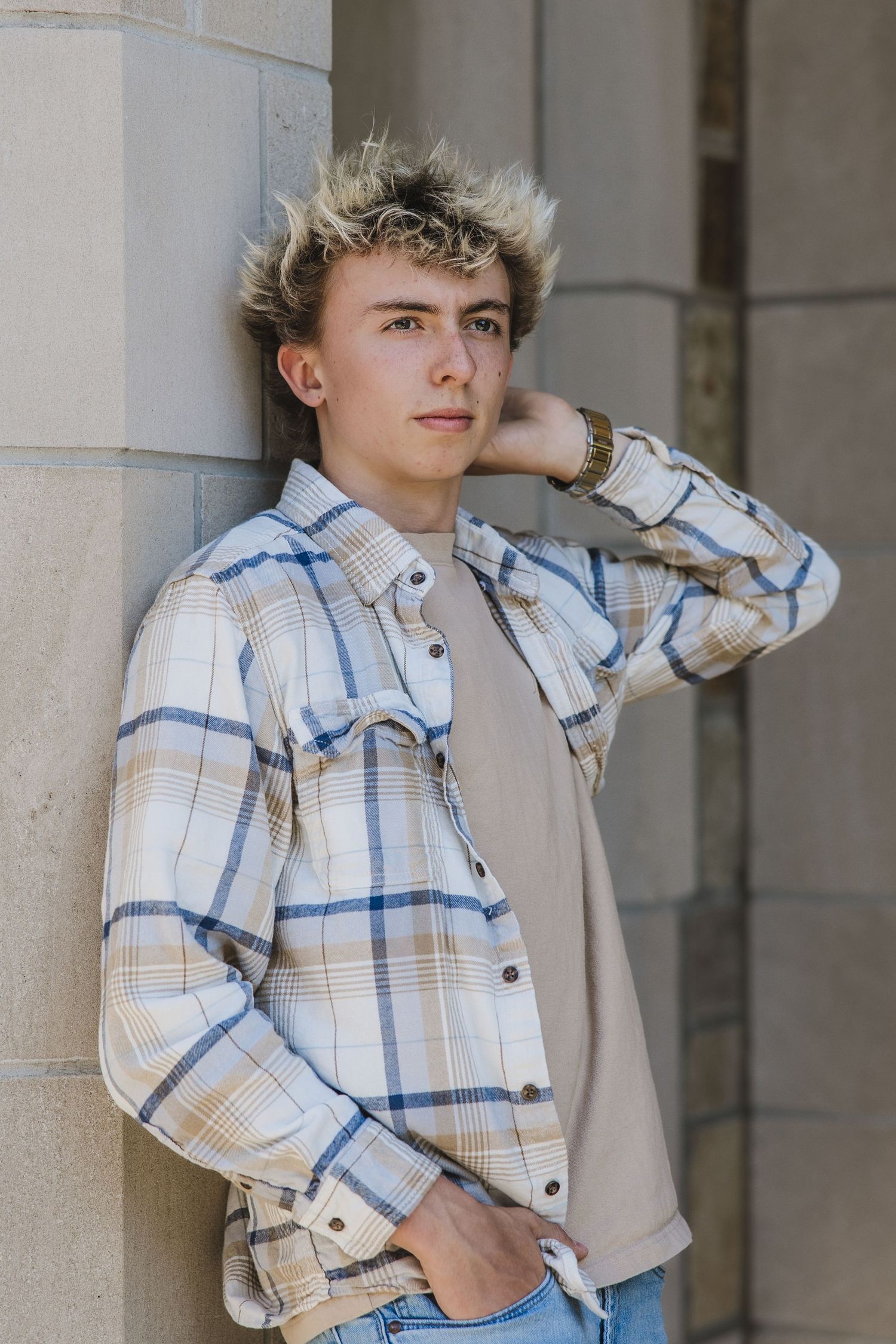 Young man in plaid shirt leaning against stone column, hand behind head, casual outdoor portrait.