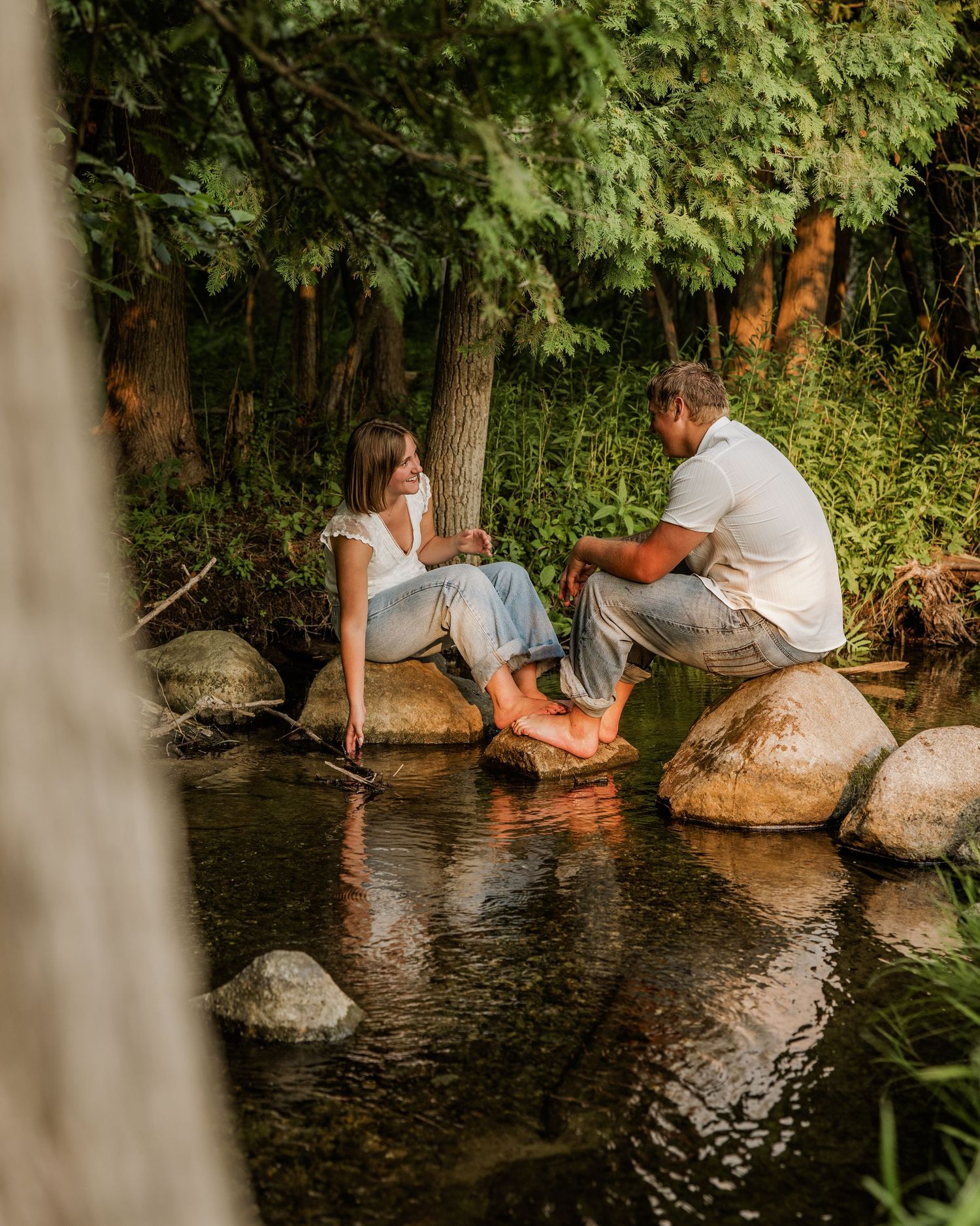 Couple sitting on rocks by a peaceful creek, barefoot and chatting, surrounded by lush green trees in a serene outdoor setting.