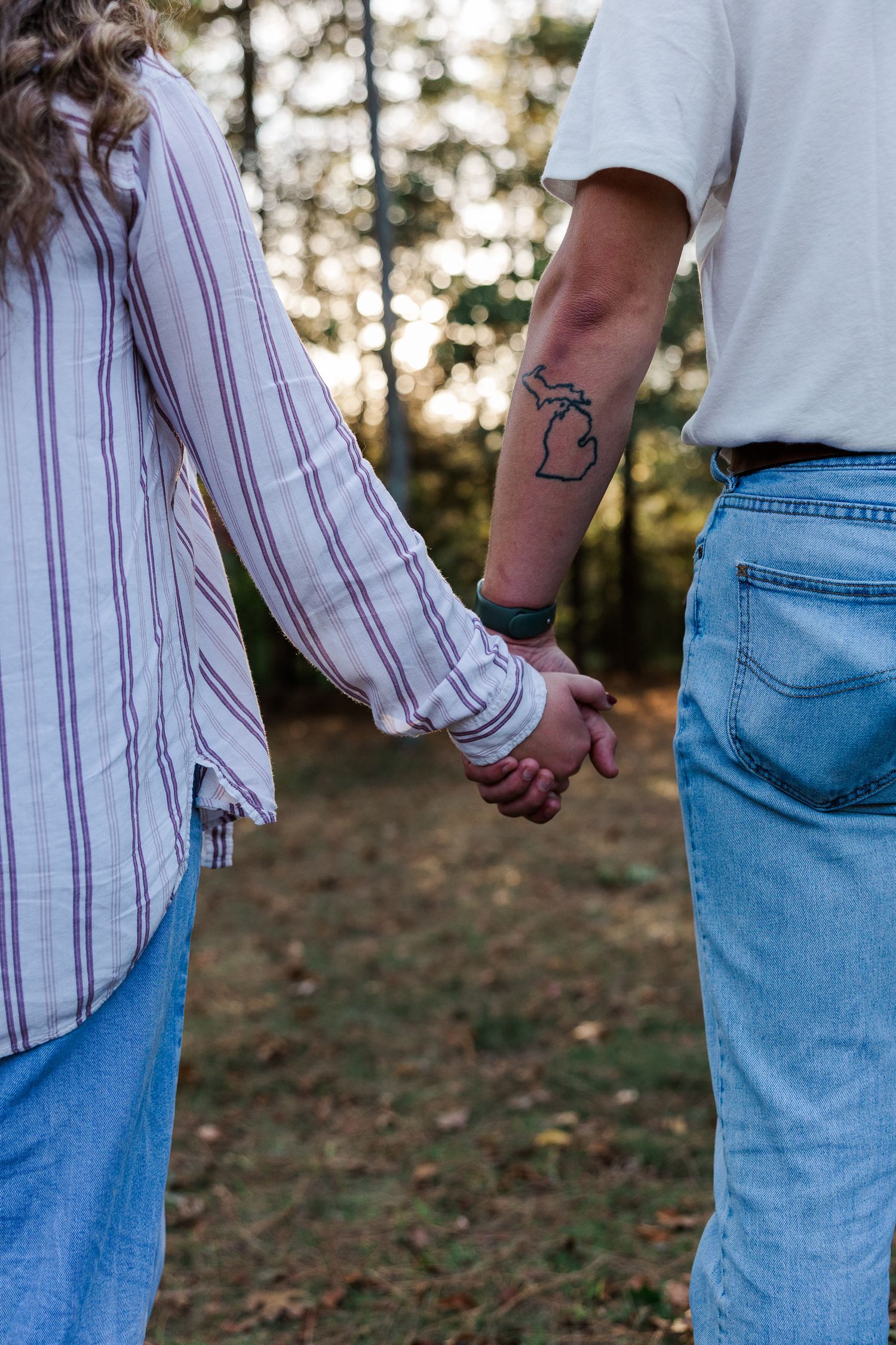 Couple holding hands in forest, man with Michigan state tattoo on forearm, wearing jeans and casual shirts.