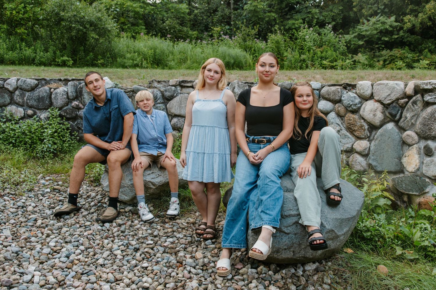 Five young people posing on rocks in front of a stone wall surrounded by lush greenery outdoors.