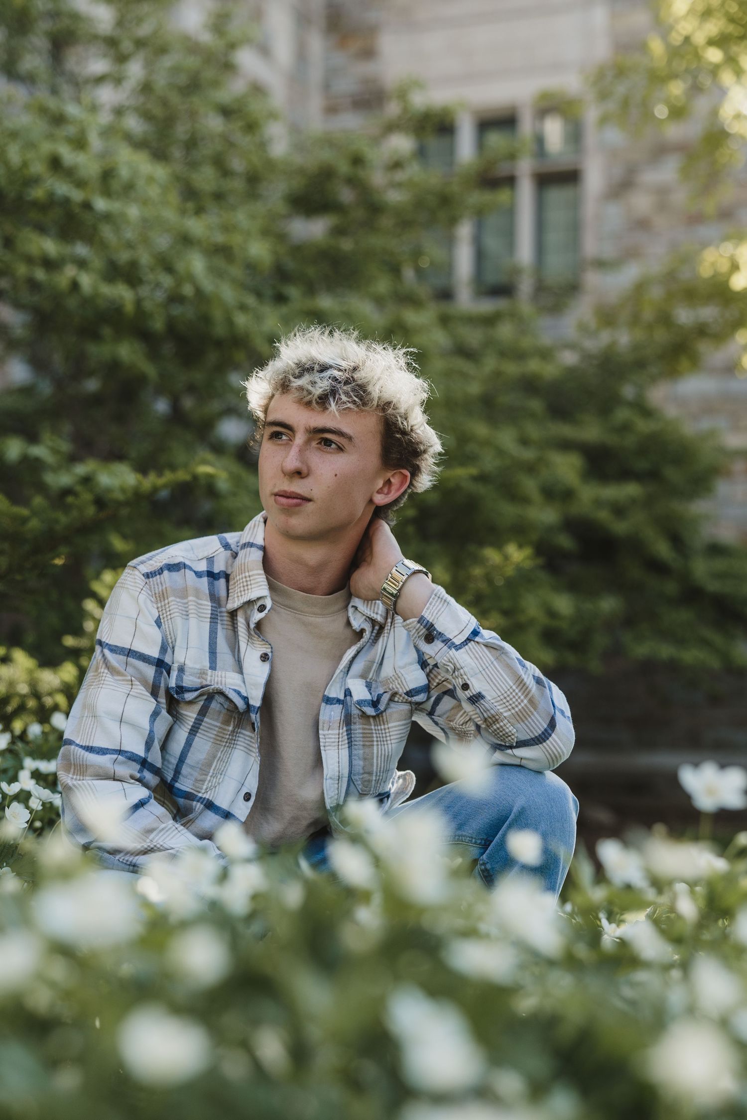 Young man with blonde curly hair sitting outdoors among white flowers, wearing plaid shirt and jeans.