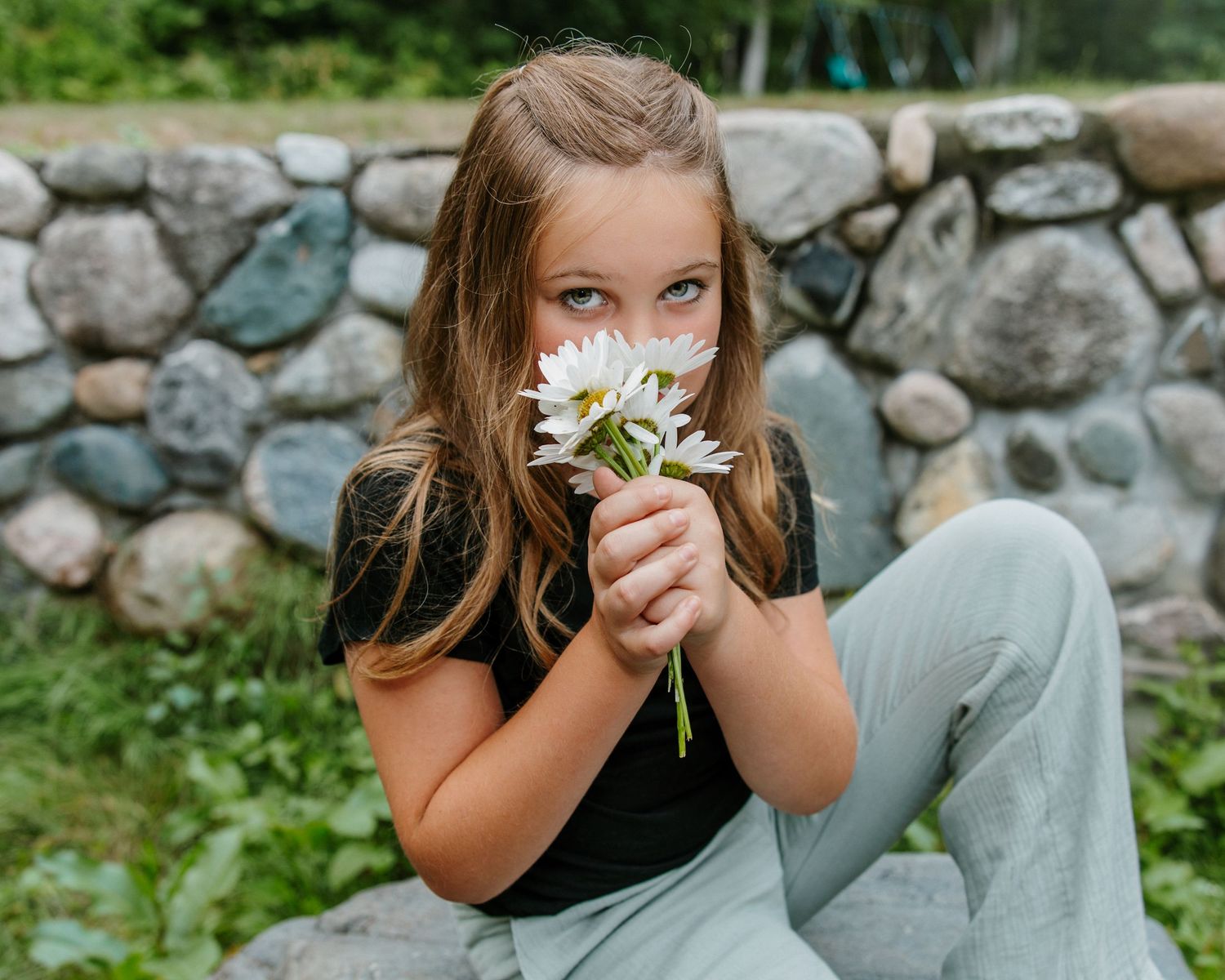 Young girl smelling white daisies in front of a rustic stone wall outdoors.