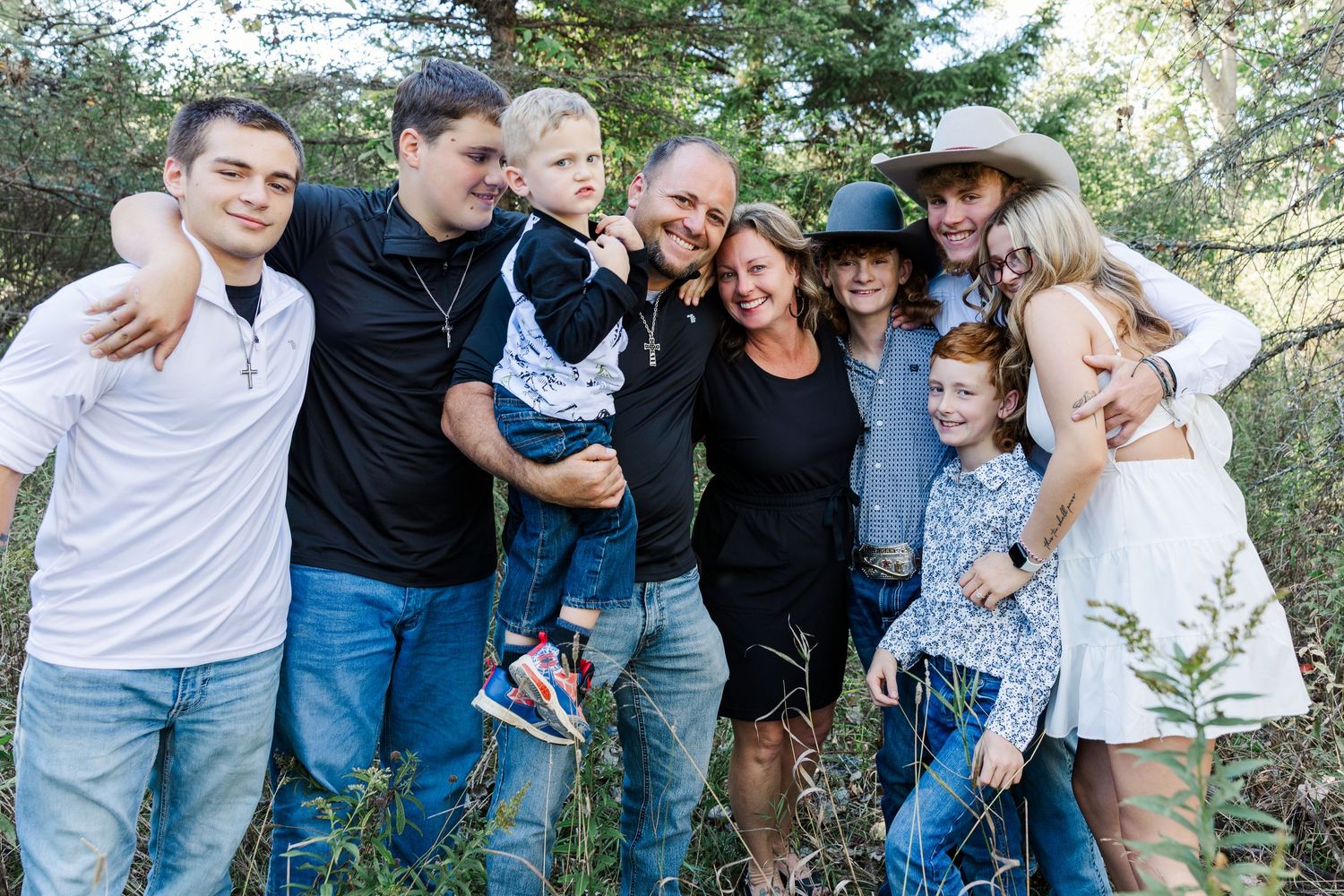 Large extended family group hugging and smiling together outdoors in a wooded setting.