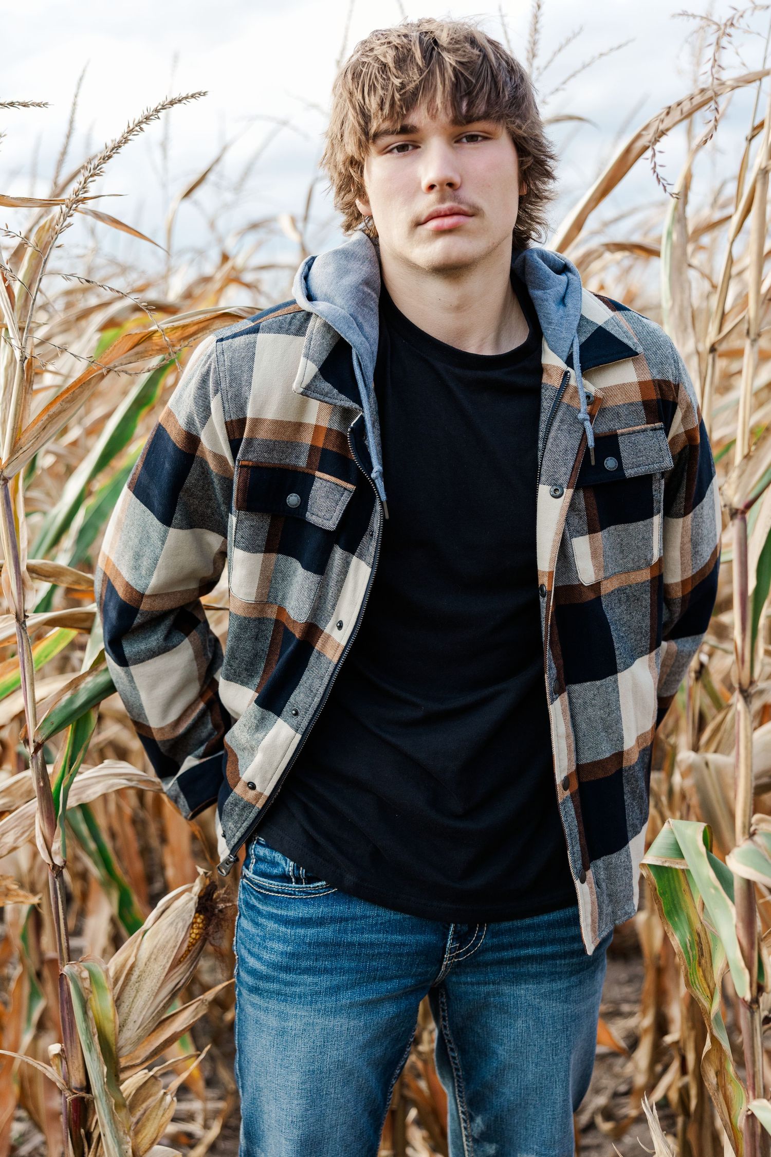 Teen boy in plaid flannel jacket standing confidently in a dried autumn cornfield.