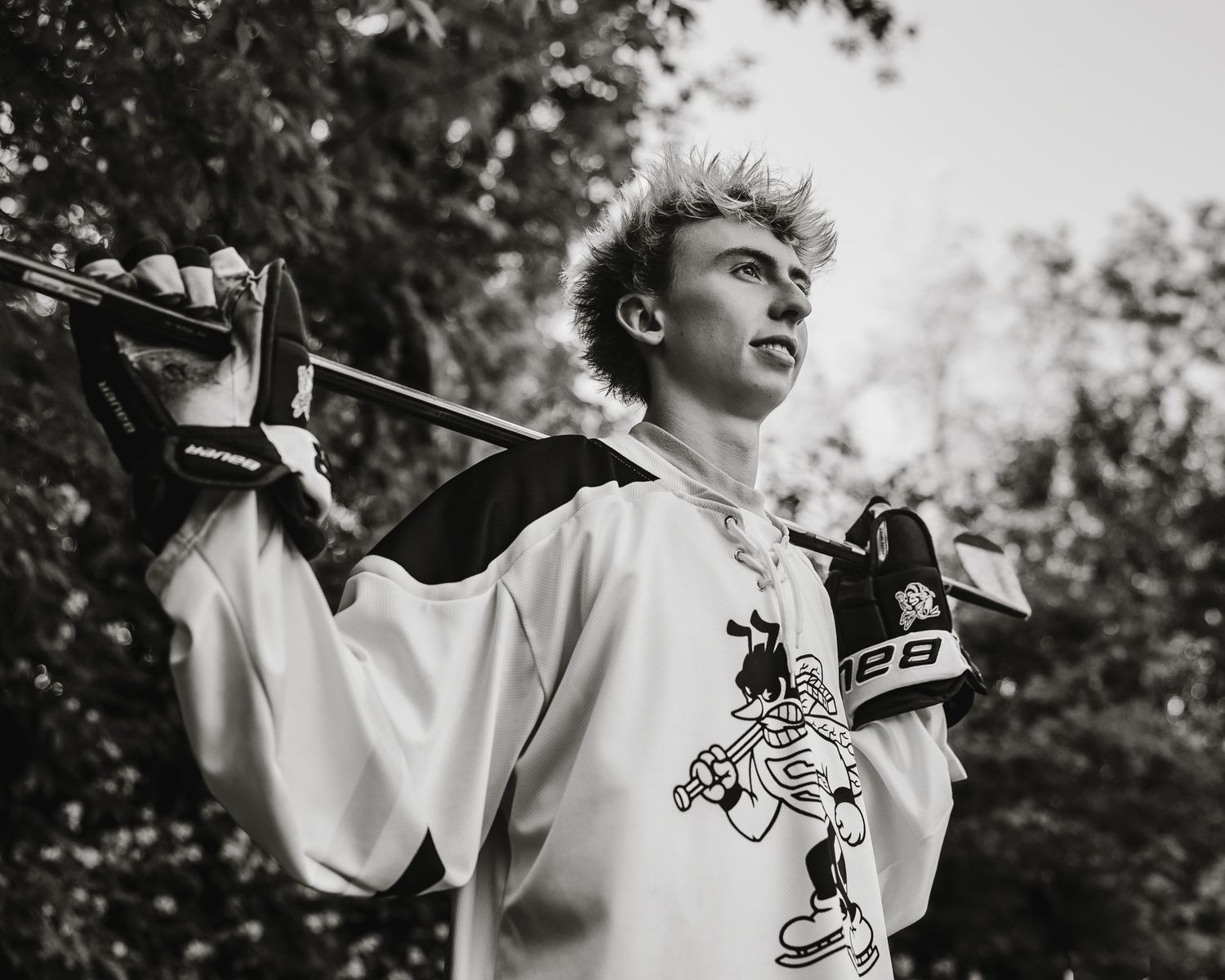 Black and white photo of a teen hockey player holding a stick over his shoulder outdoors.