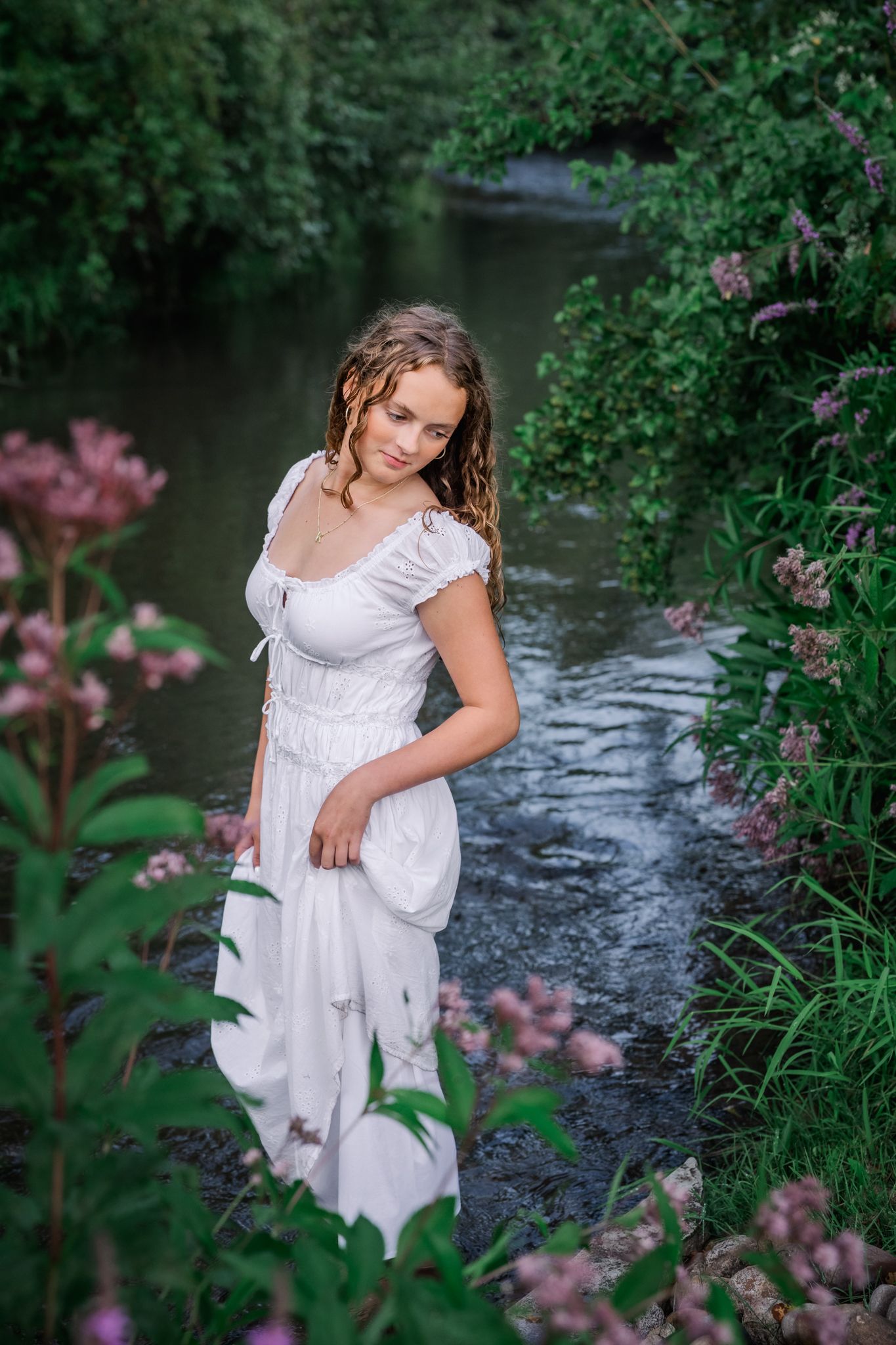 Young woman in white dress standing in a serene stream surrounded by lush greenery and pink wildflowers.