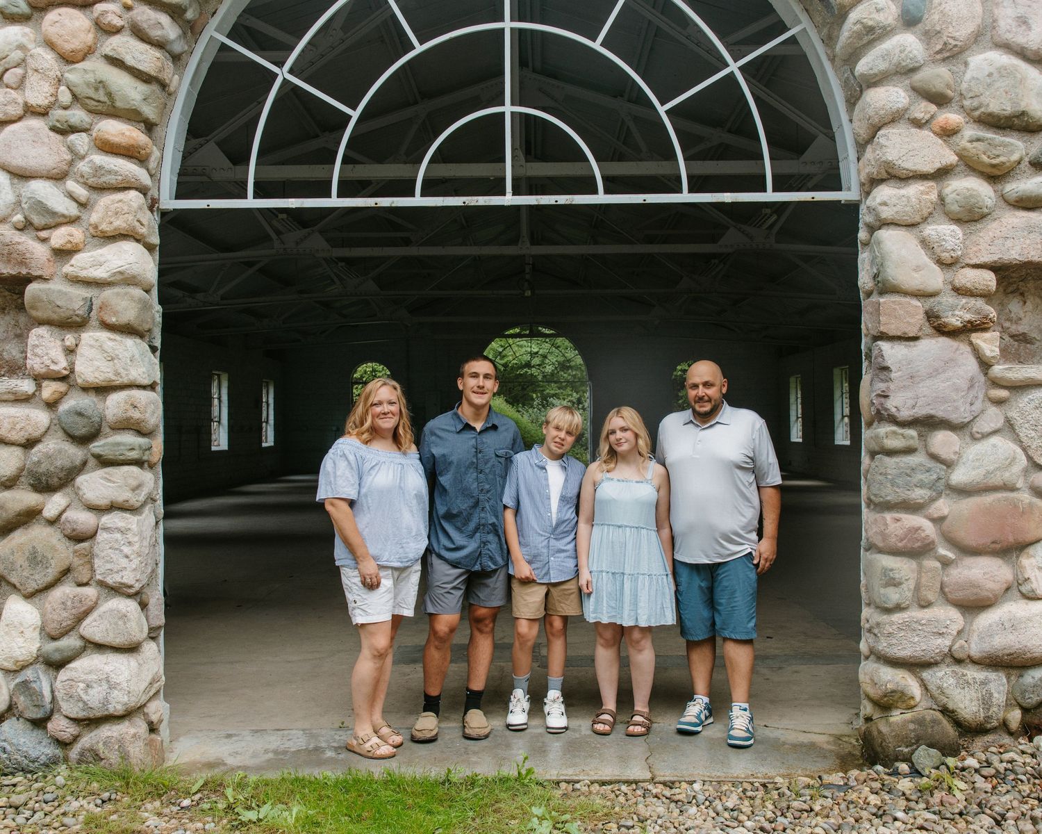 Family of five posing under a stone arched entrance, smiling together outdoors on a summer day.