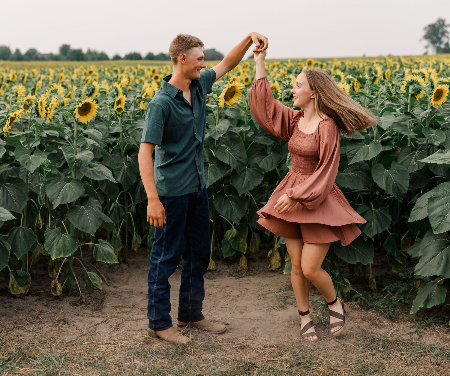 Couple dancing joyfully in a sunflower field, man spinning woman in rust dress, romantic outdoor portrait.