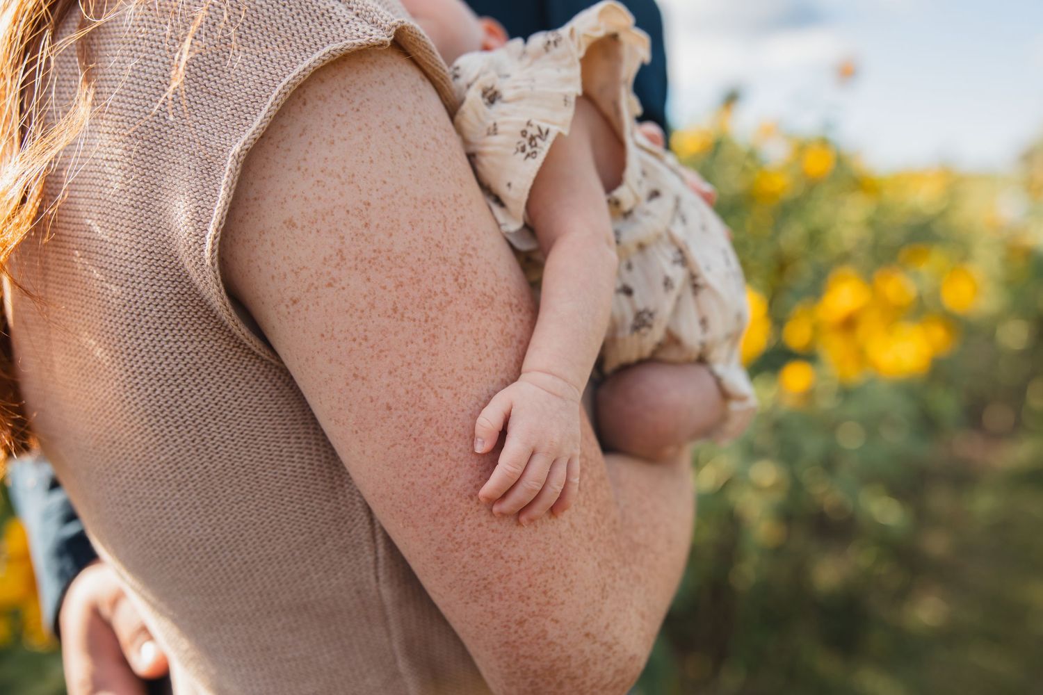 A freckled mother holds her baby in a floral outfit among sunflowers in a warm outdoor setting.