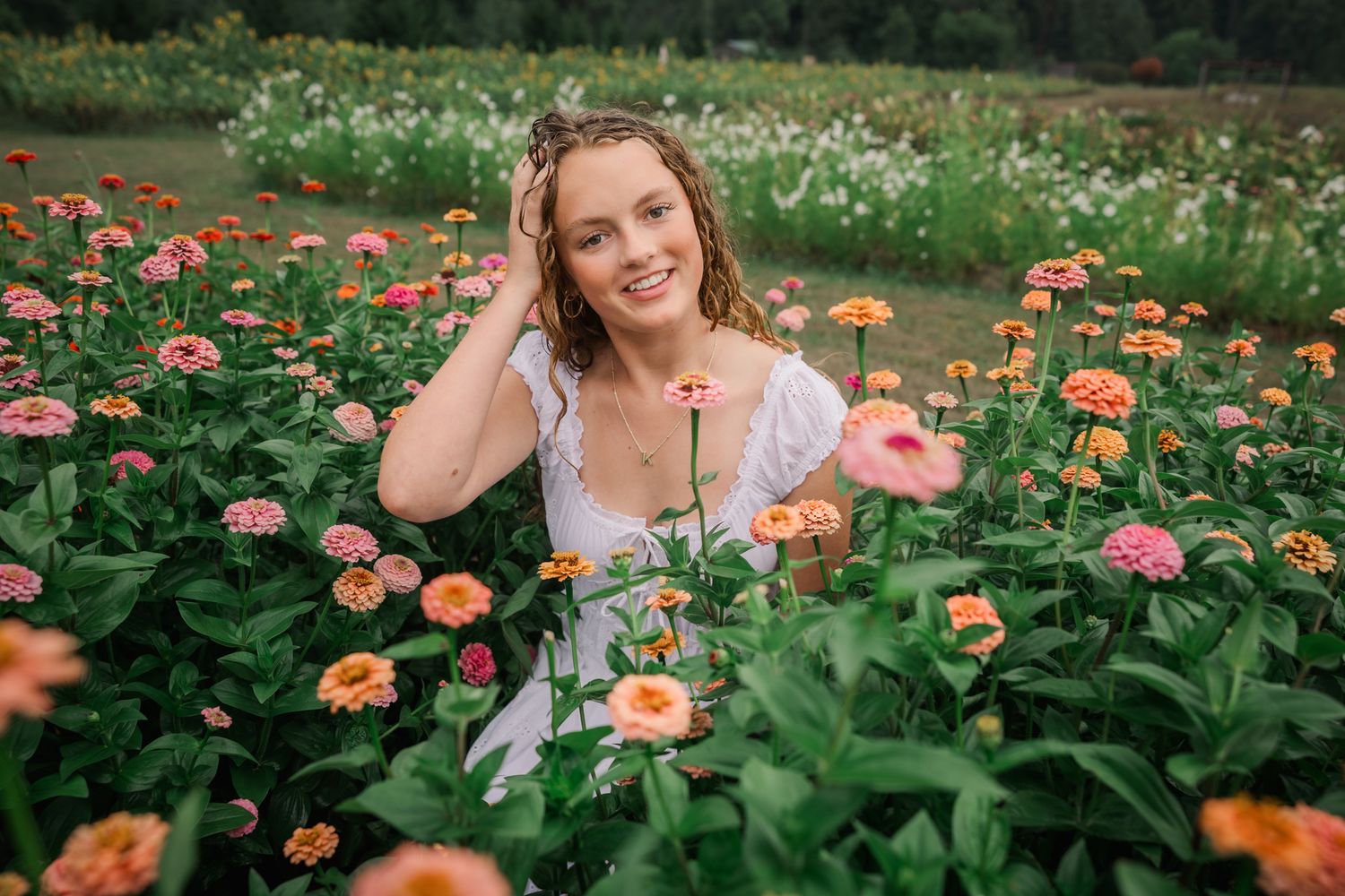 Young woman in white dress smiling among colorful pink and orange zinnia flowers in a lush garden field.