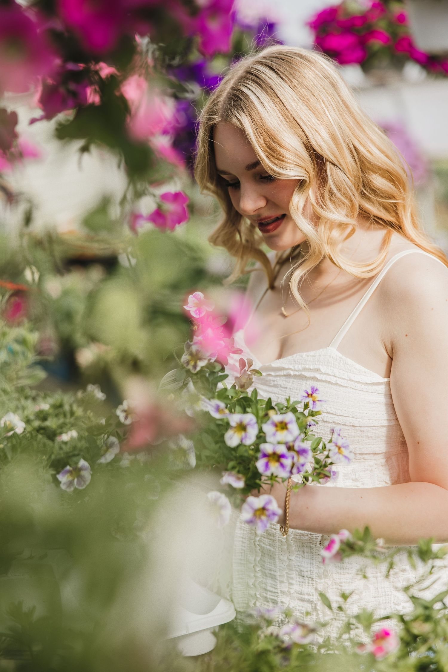 Blonde woman in white dress admiring pink and purple flowers in a lush garden setting.