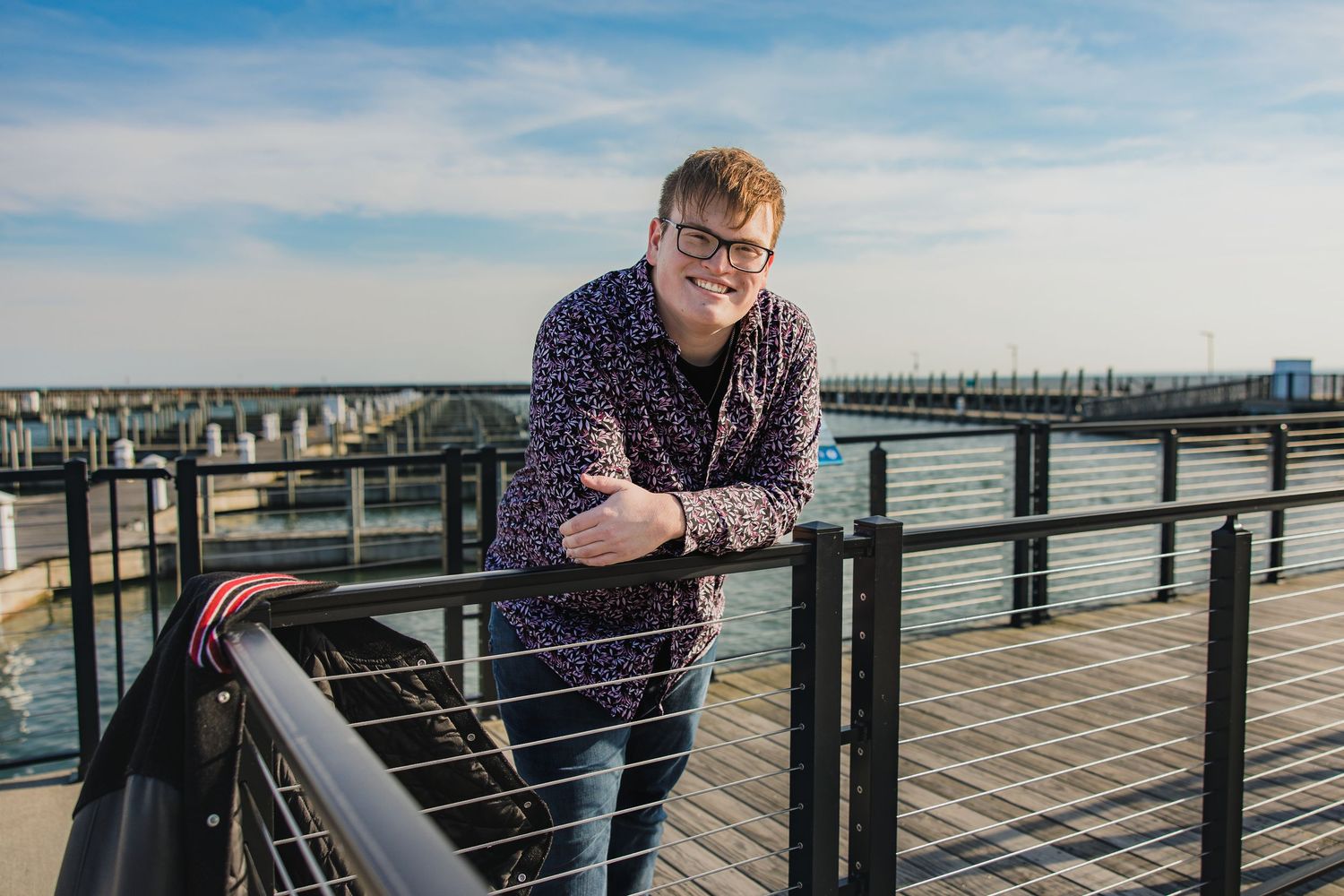 Young man with glasses leaning on a railing at a waterfront marina on a sunny day.