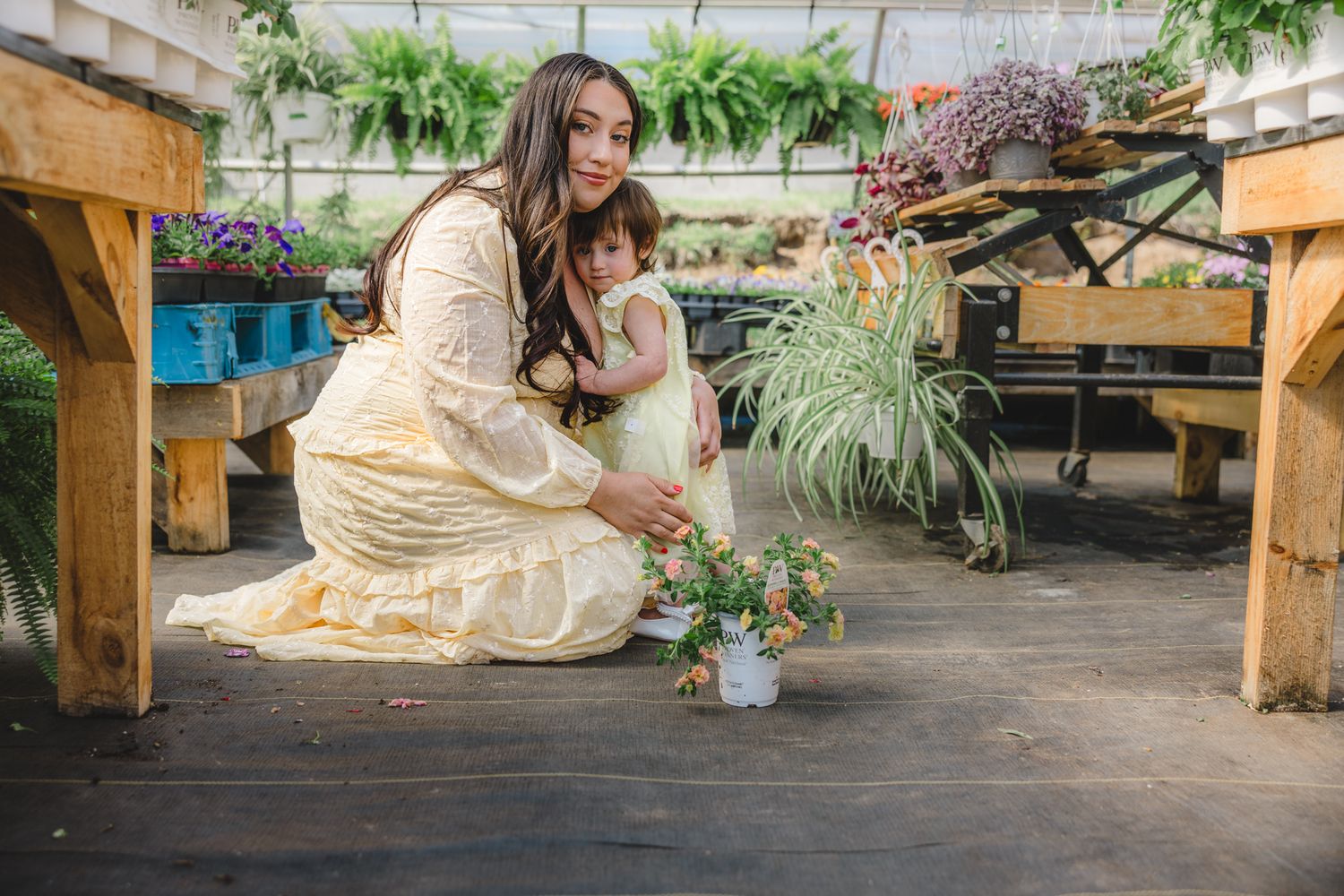 A mother and young daughter sit together on a greenhouse floor surrounded by lush plants and flowers.