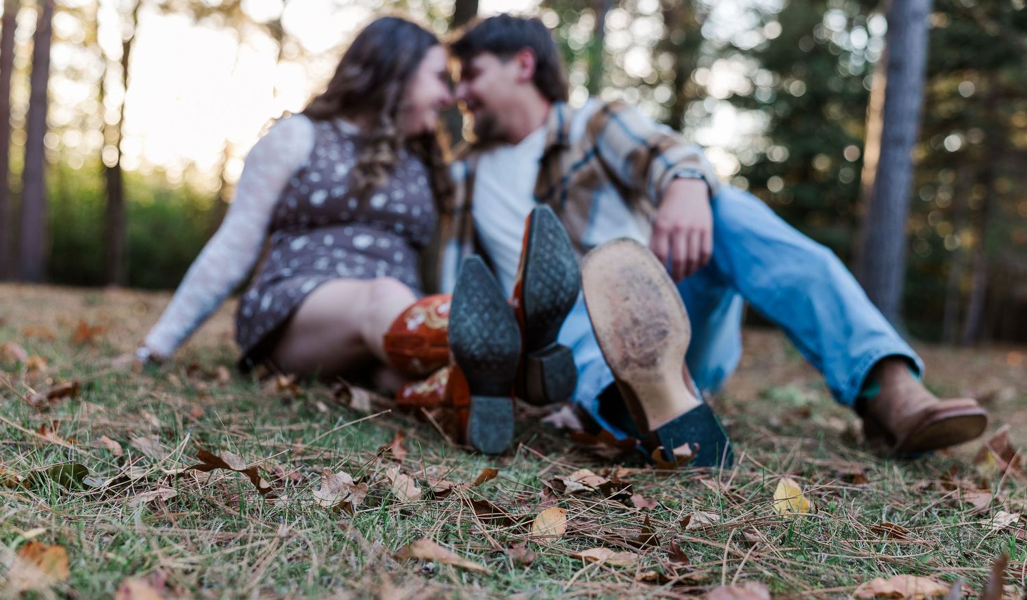 Couple sitting on autumn leaves in forest, boots in foreground, leaning close together romantically.