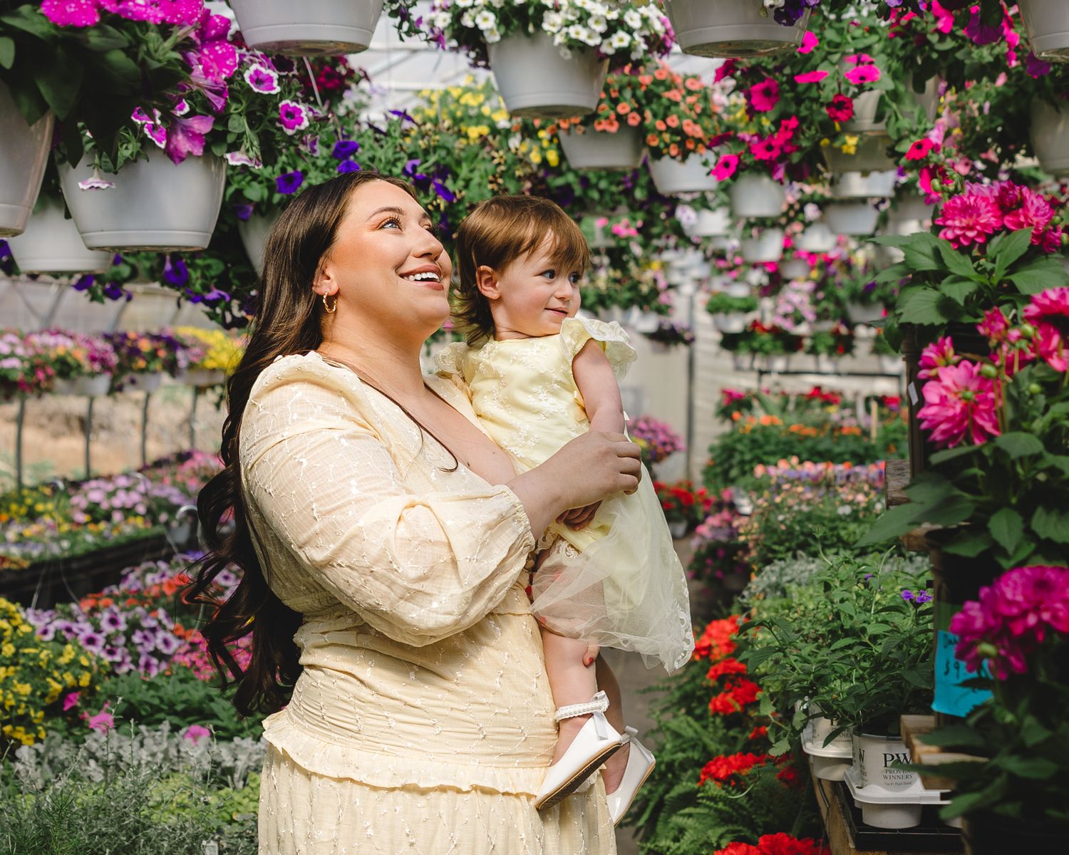 Smiling mother holding laughing toddler daughter in yellow dresses inside colorful flower greenhouse.