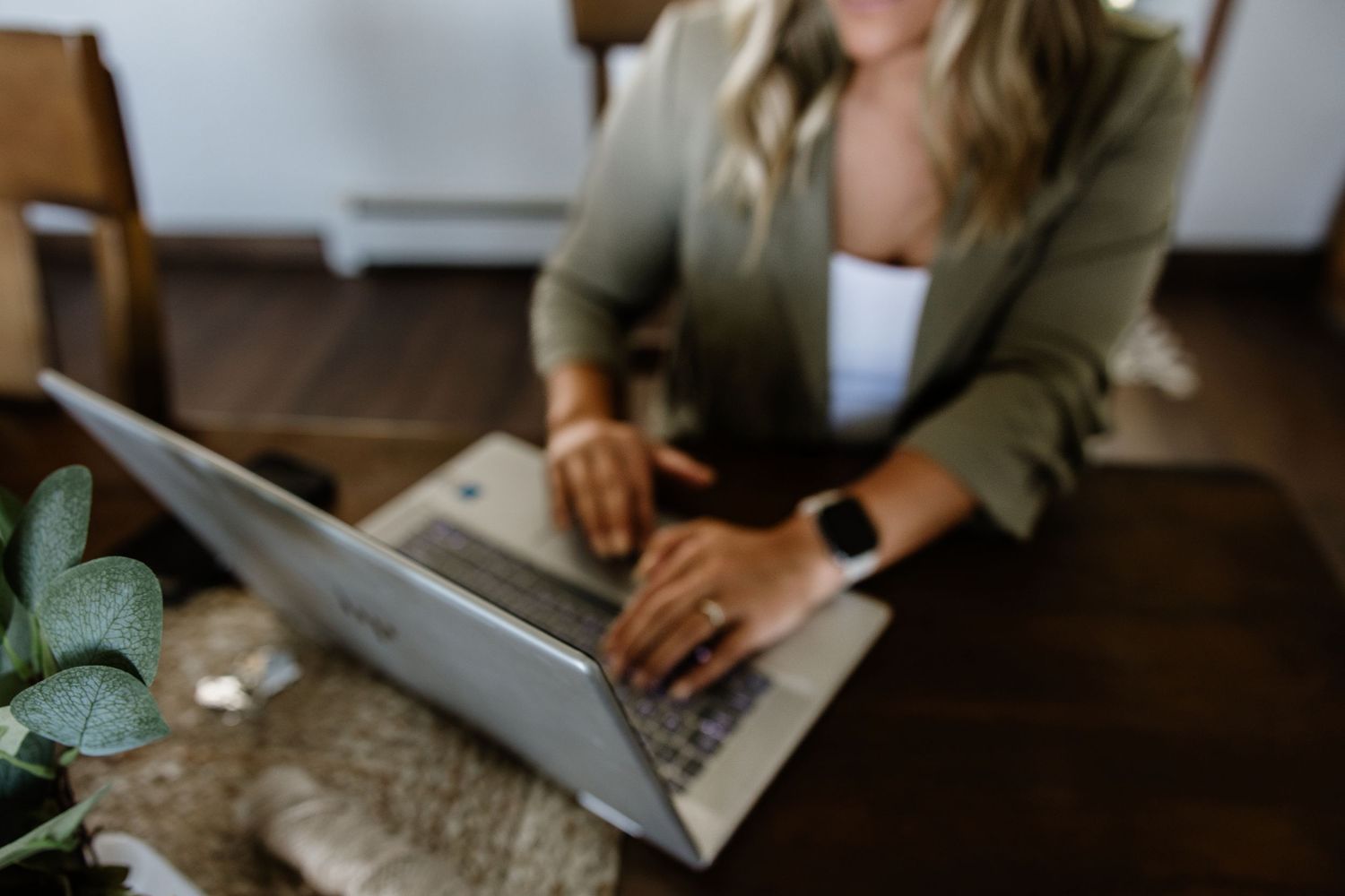 Blonde woman in olive blazer typing on laptop at wooden desk with plant in foreground.