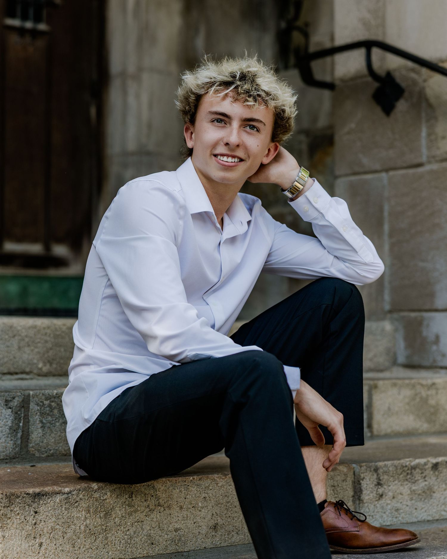 Young man in white shirt and dark pants sitting on stone steps, smiling confidently outdoors.