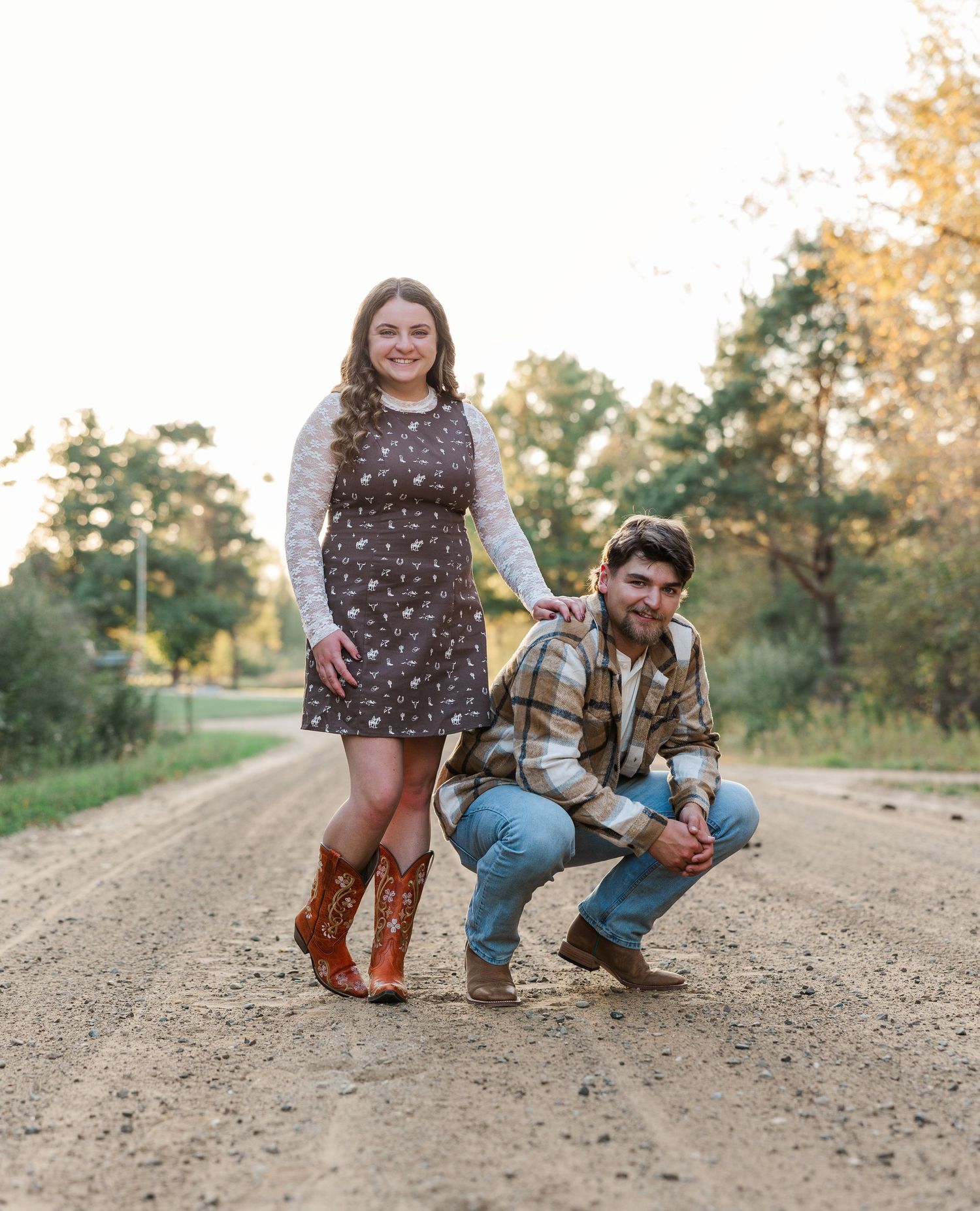 Couple posing on a dirt road, woman in floral dress and cowboy boots, man crouching in plaid shirt and jeans.