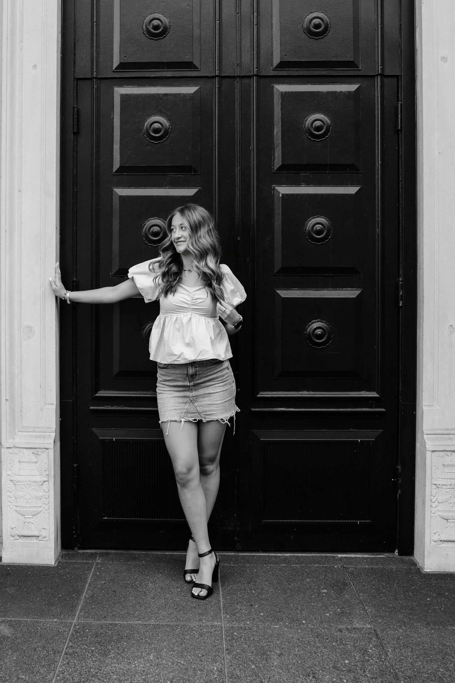 Black and white photo of a young woman in a peplum top and denim skirt posing by large ornate wooden doors.