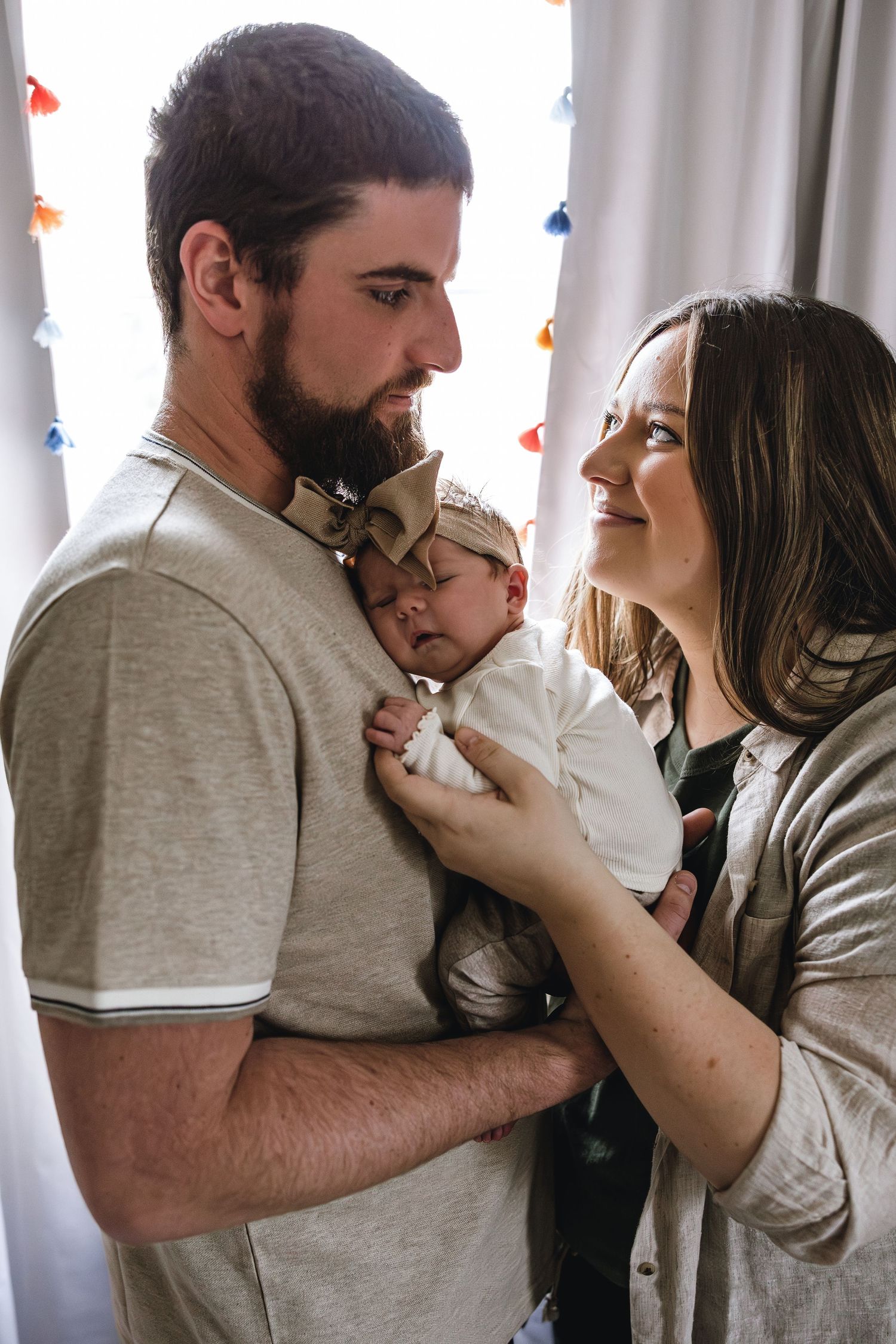 A bearded man and smiling woman lovingly hold their newborn baby by a bright window.
