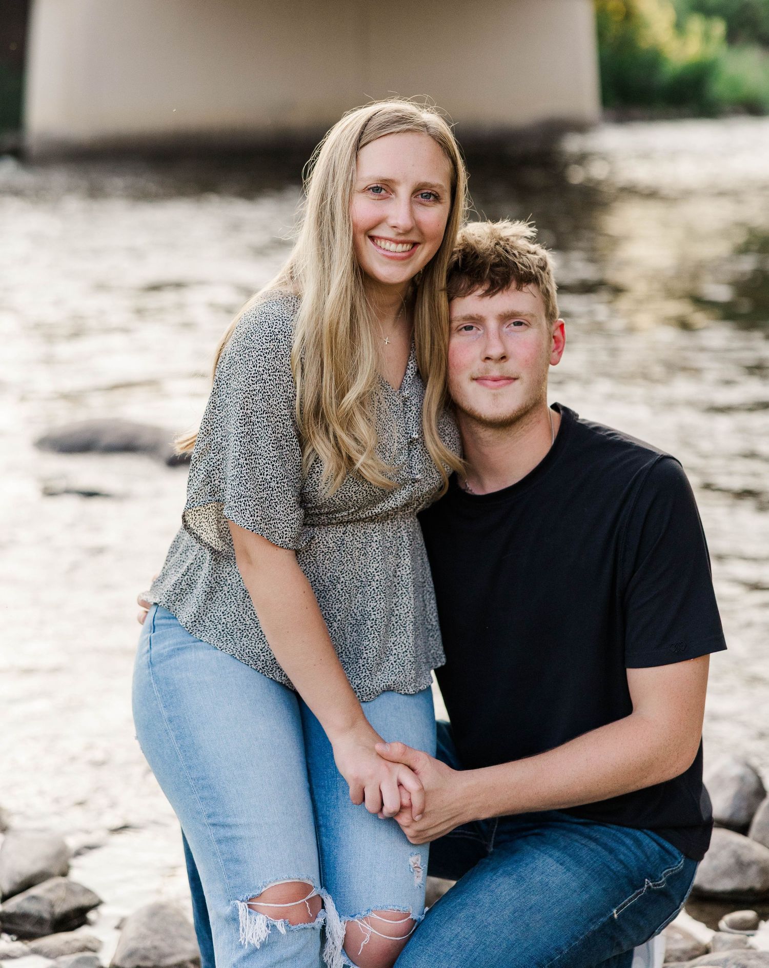 A young couple smiling and posing together near a rocky riverbank with a bridge in the background.