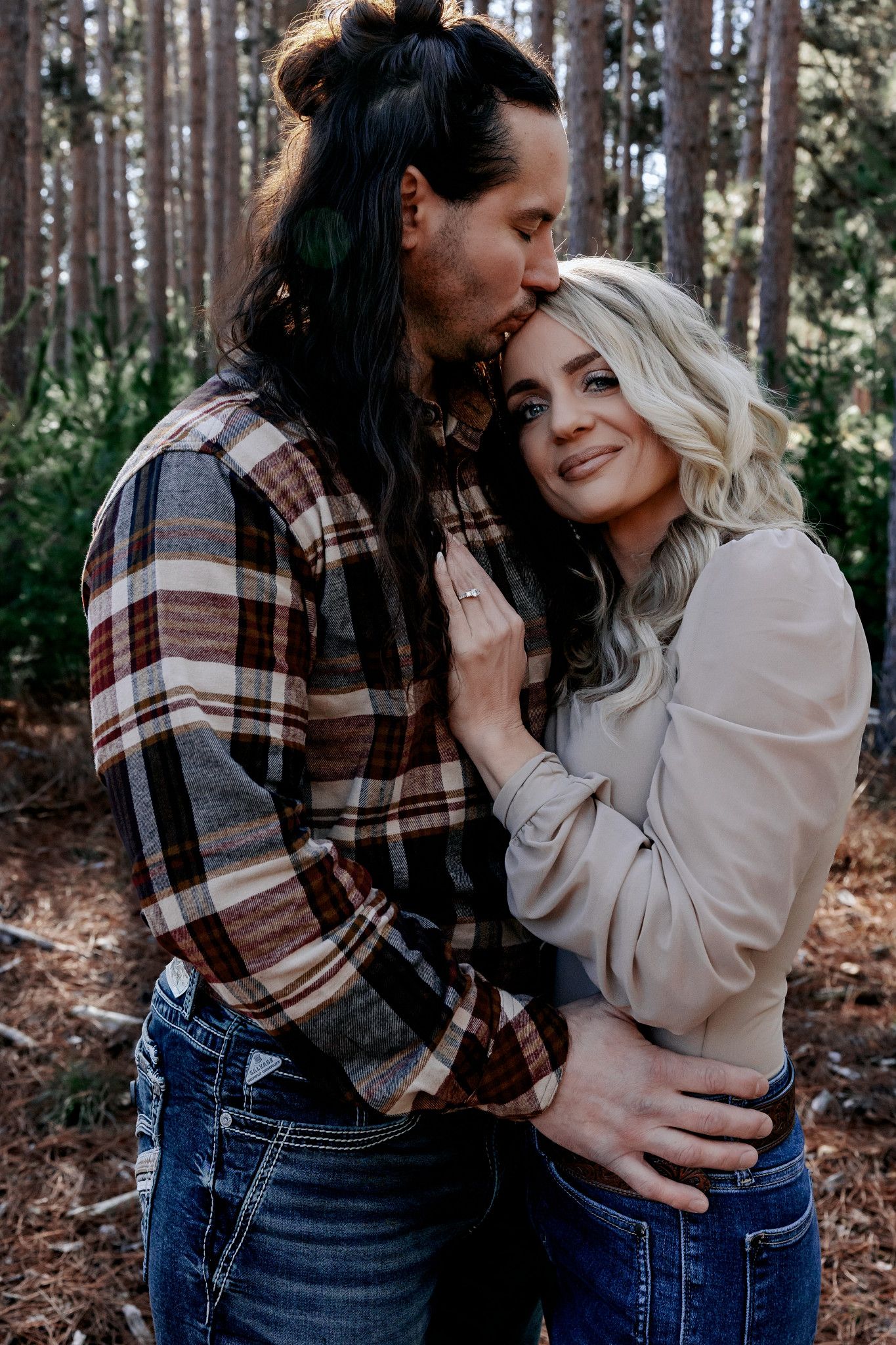 Couple embracing in a pine forest, man kissing womans temple, wearing flannel and casual tops with jeans.