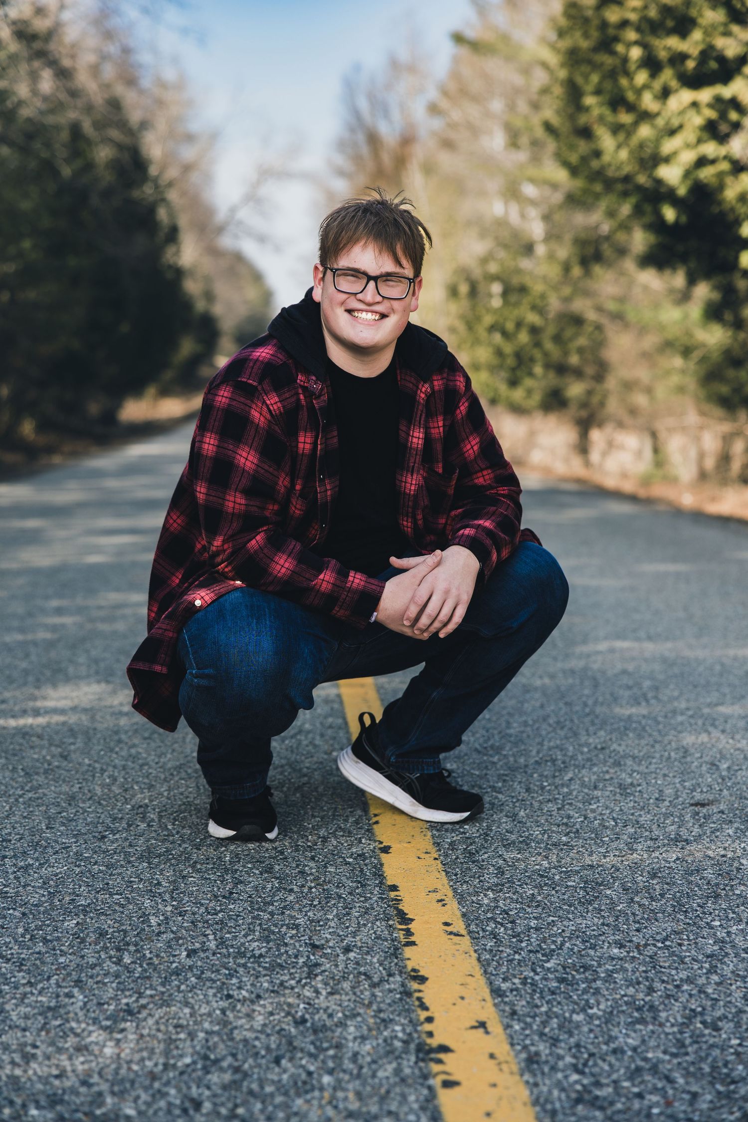 Teen boy in red plaid shirt and jeans crouching on a road centerline, smiling, wearing glasses, outdoors.