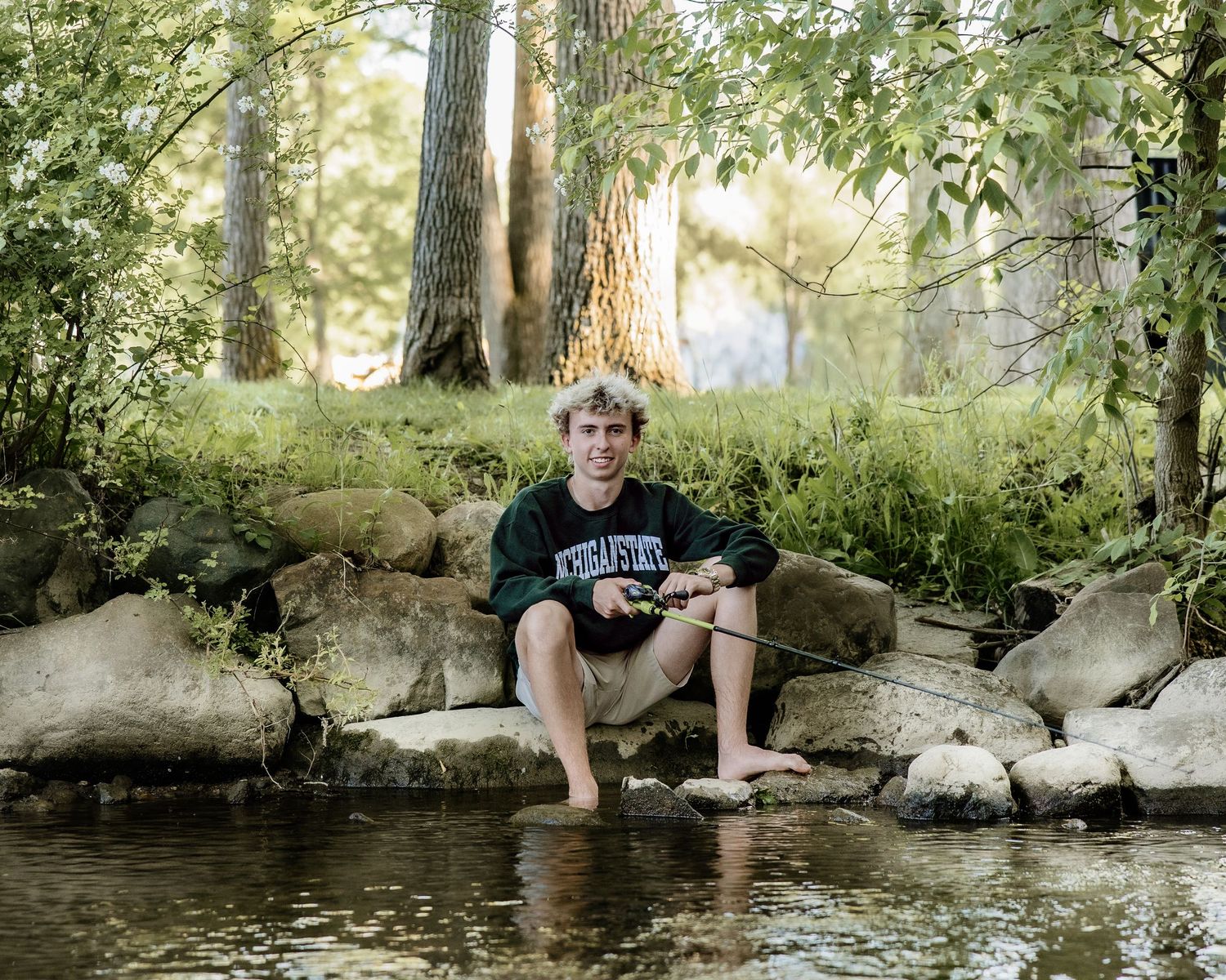 Young man sitting barefoot on rocks fishing in a serene woodland stream surrounded by trees.