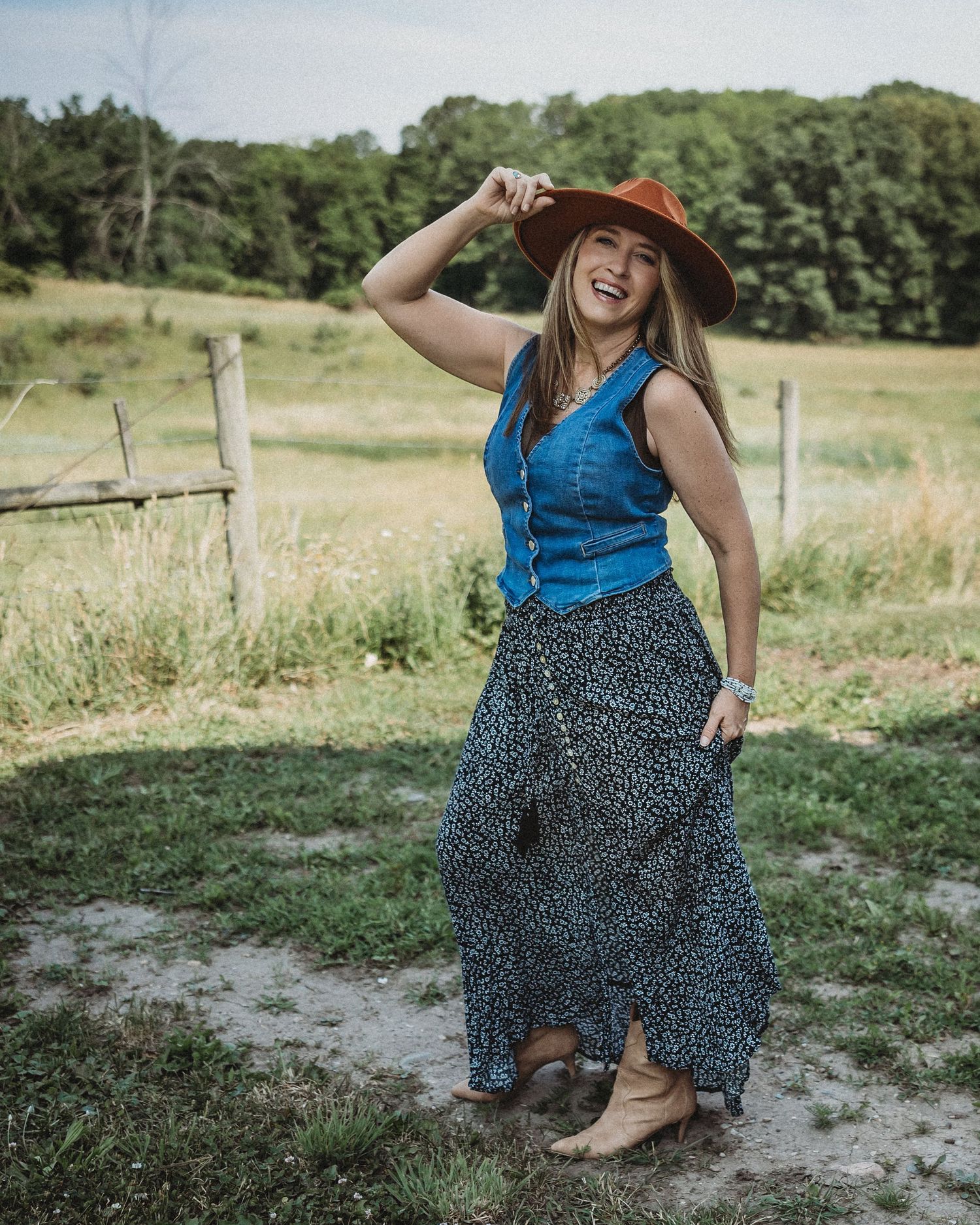 Smiling woman in denim vest, floral maxi skirt, and brown wide-brim hat posing on a rural farm path.