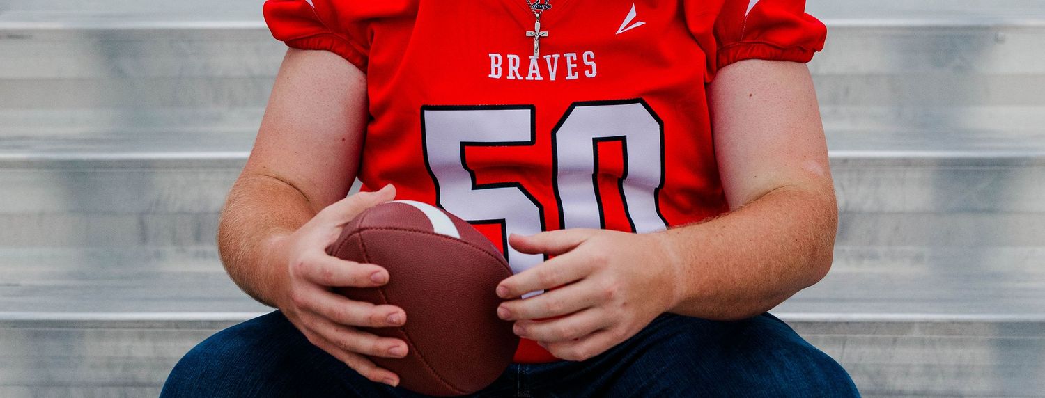 Young football player in red Braves jersey number 50 holding a football, seated on bleachers.