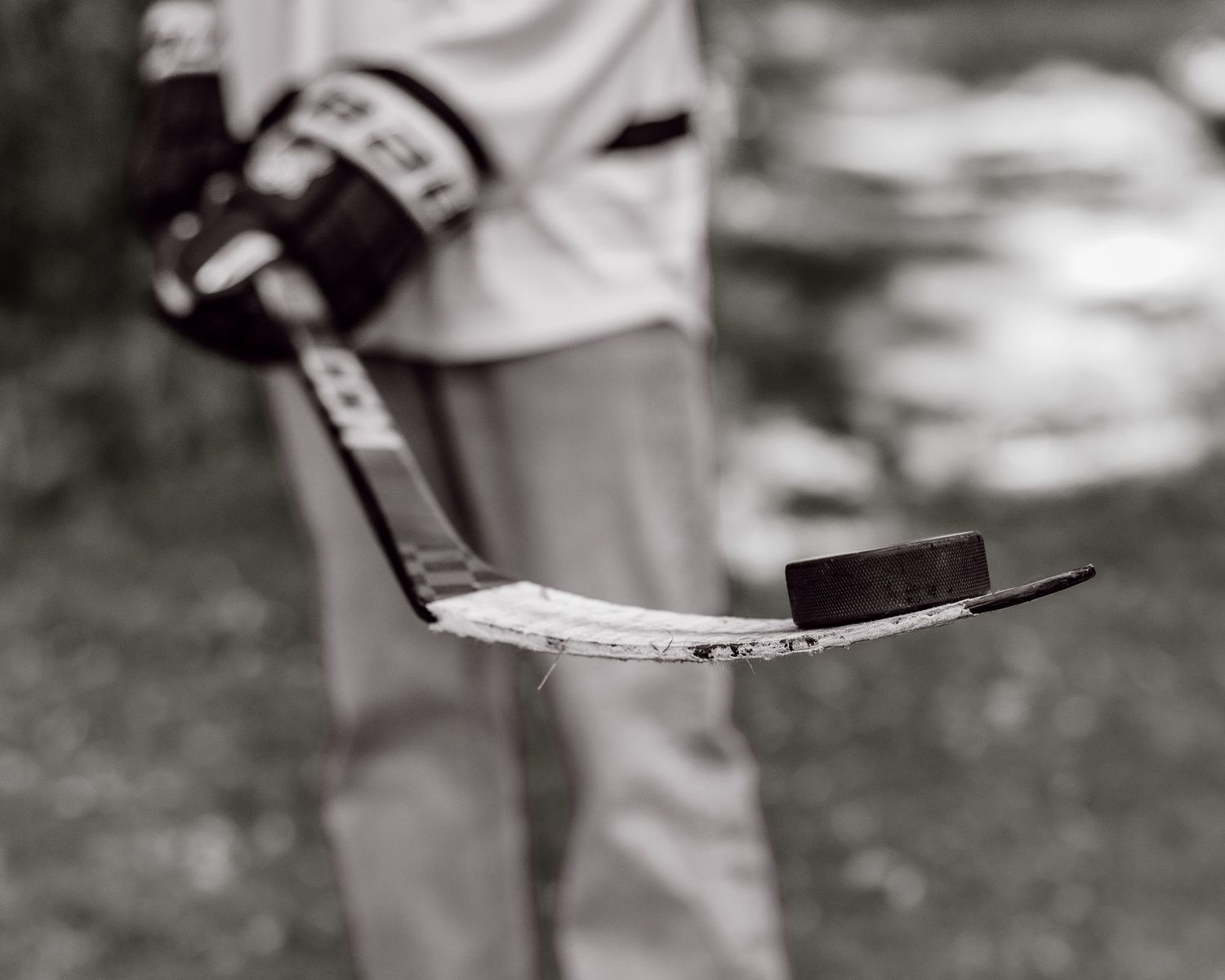 Black and white photo of hockey player holding stick with puck balanced on blade outdoors.