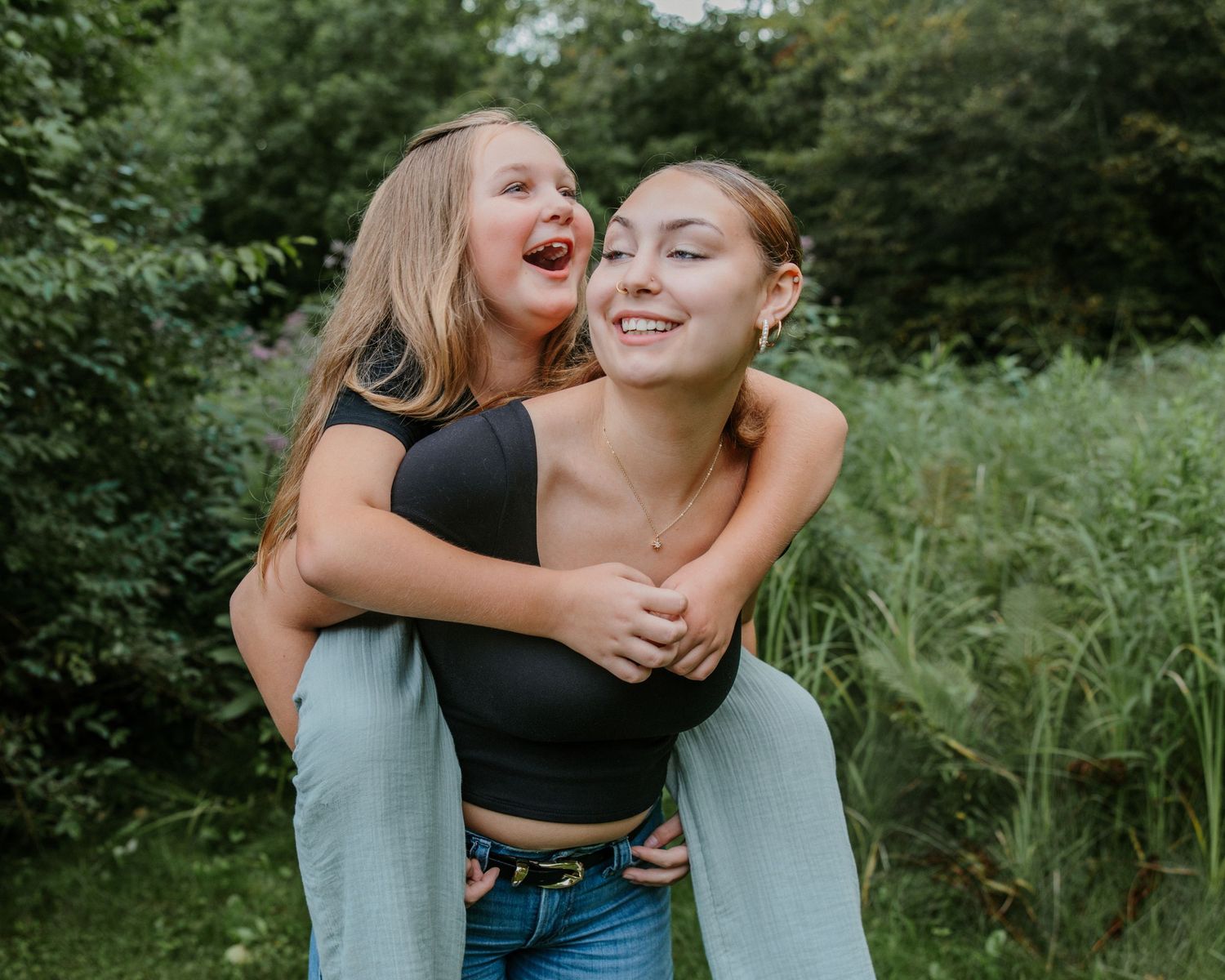 Two girls laugh outdoors, one giving the other a piggyback ride near lush green trees.