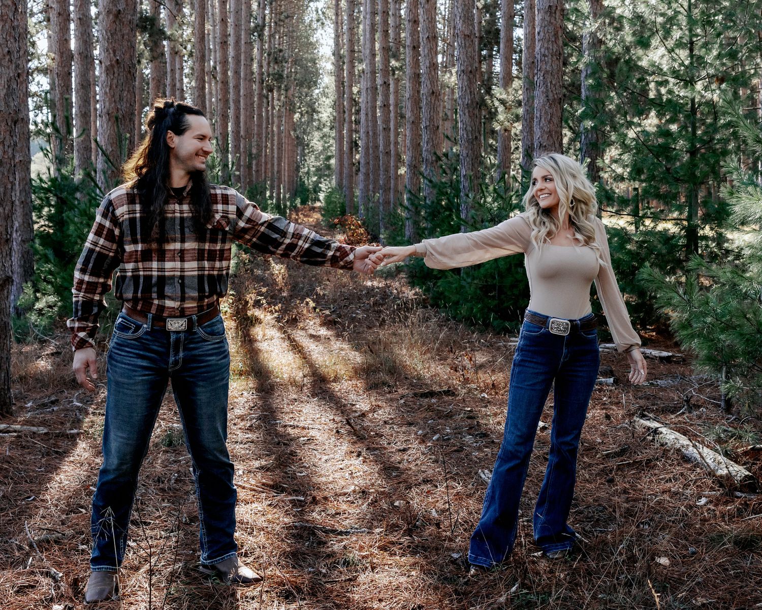Couple holding hands and dancing in a sunlit pine forest, wearing flannel and jeans in a romantic outdoor engagement photo.