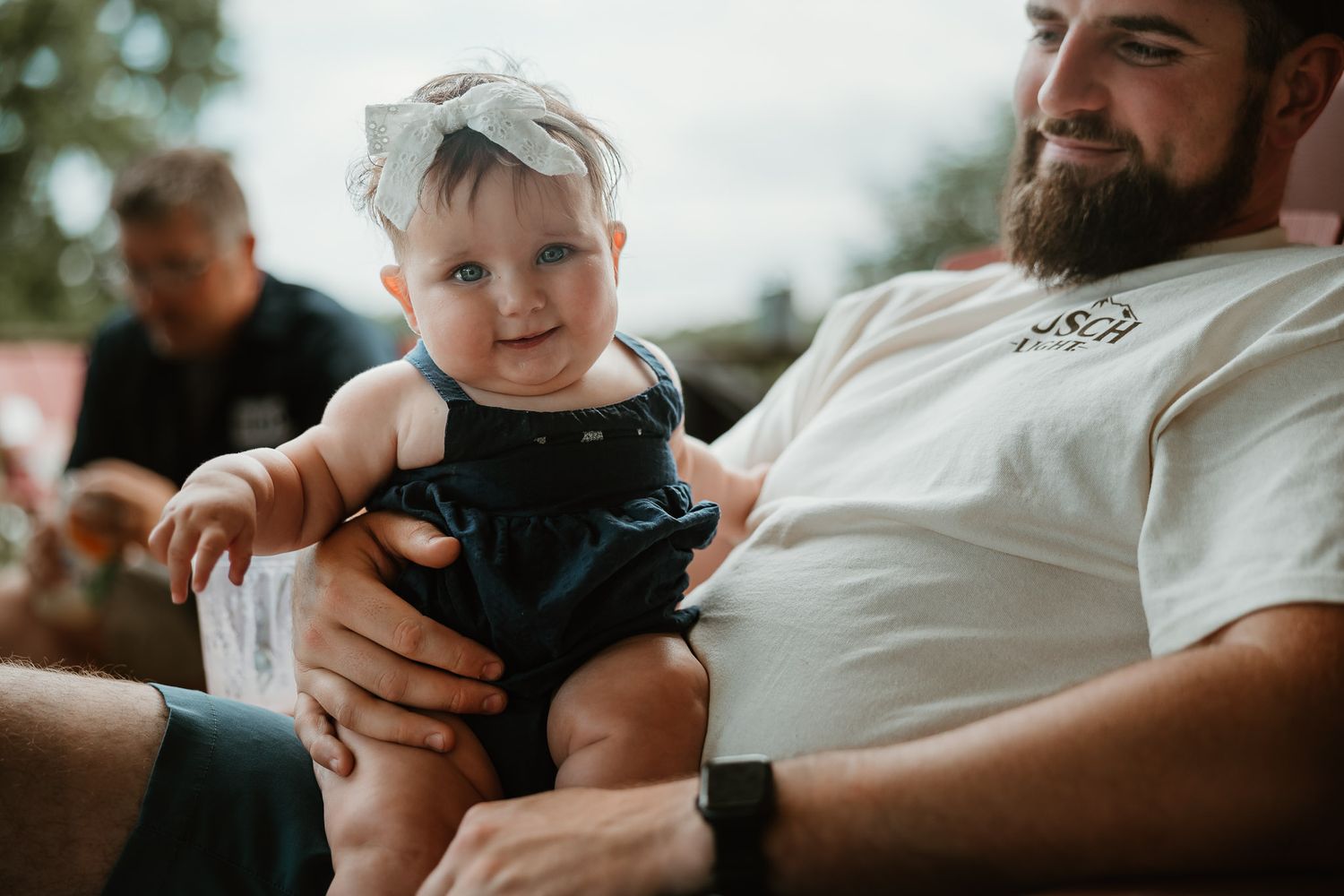 Smiling baby in navy romper being held by a bearded man in a white shirt outdoors.