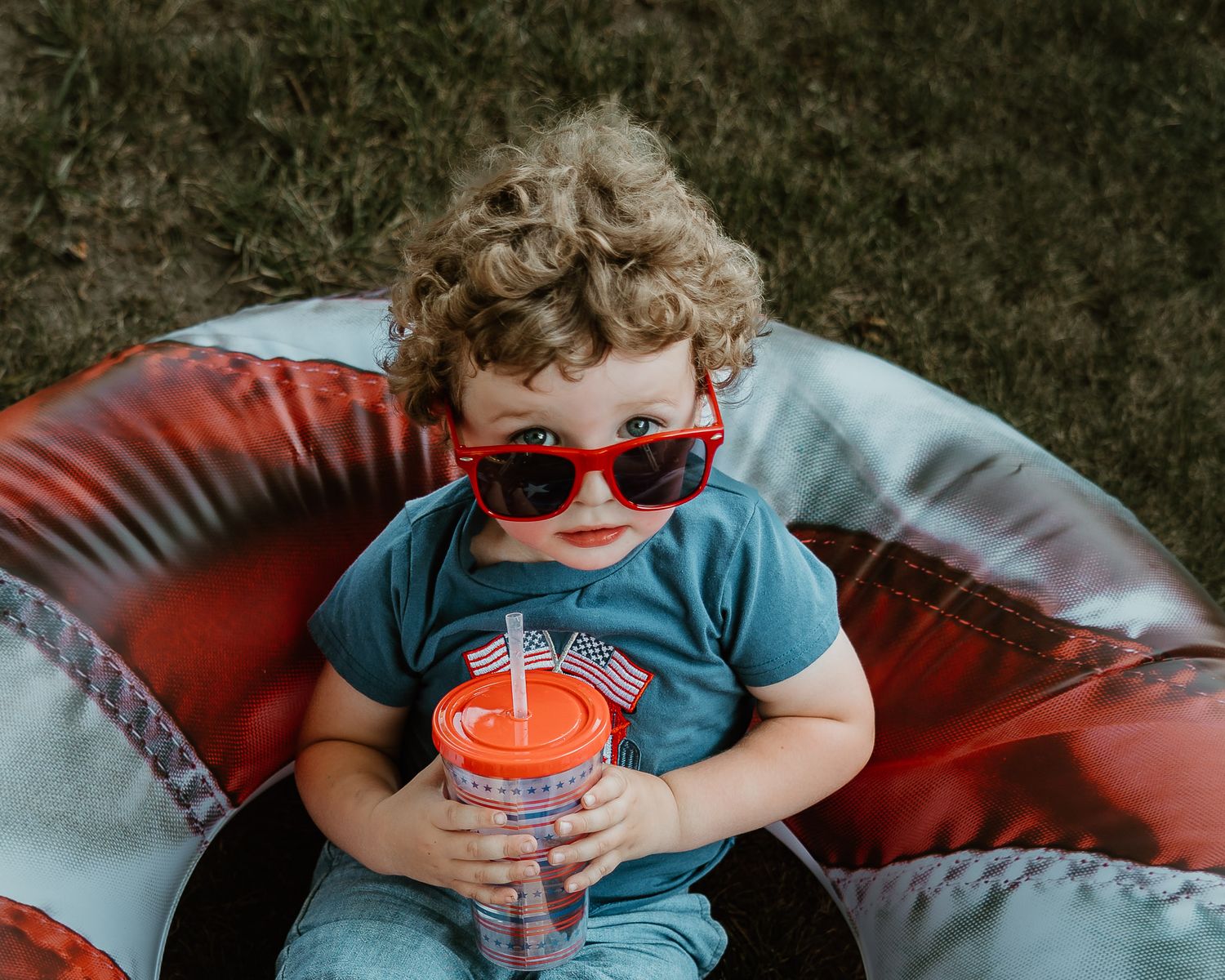 Curly-haired toddler wearing red sunglasses holding an American flag cup on a bean bag chair.