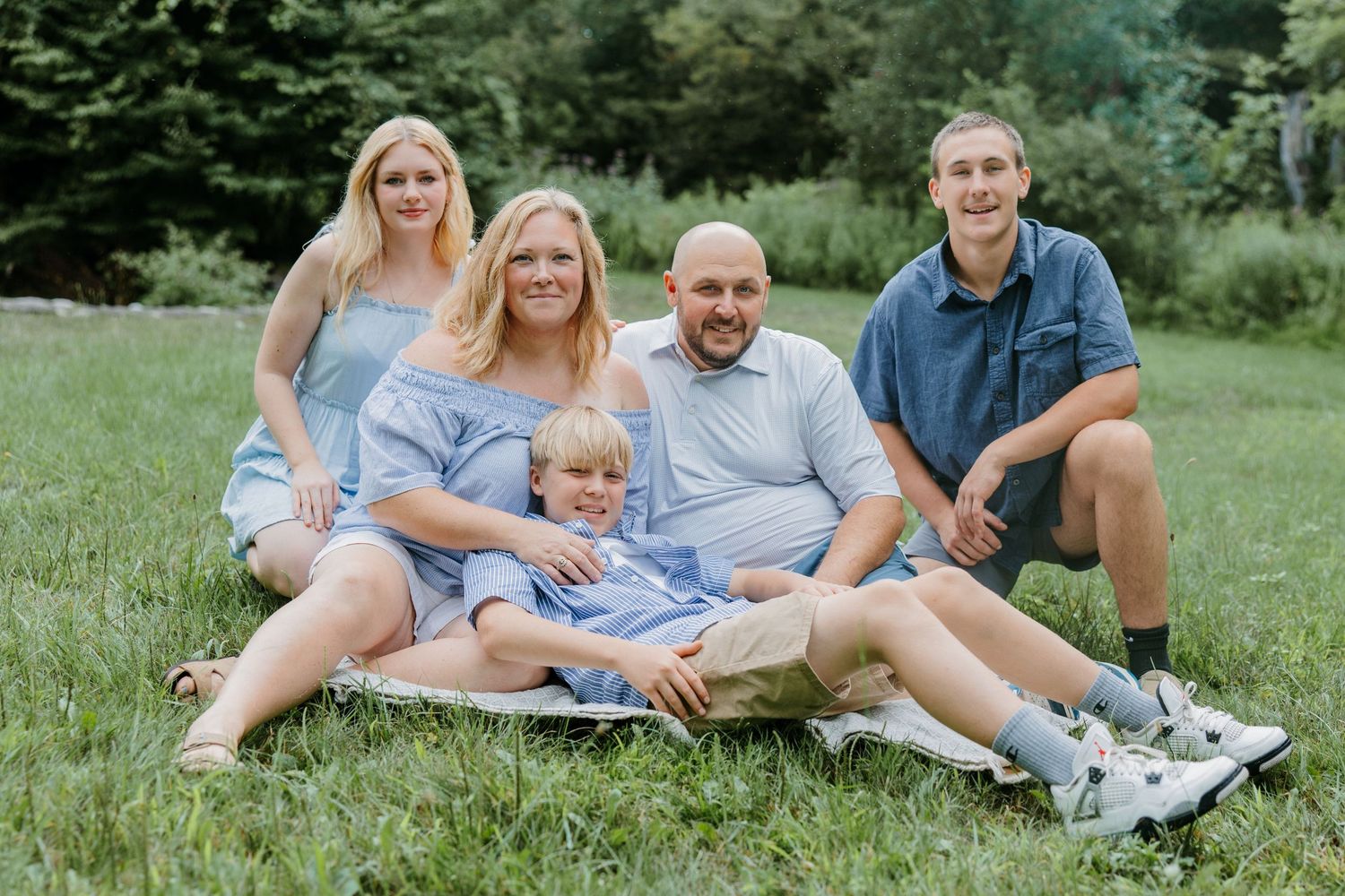 Family of five smiling and posing together on green grass with lush trees in the background on a sunny day.