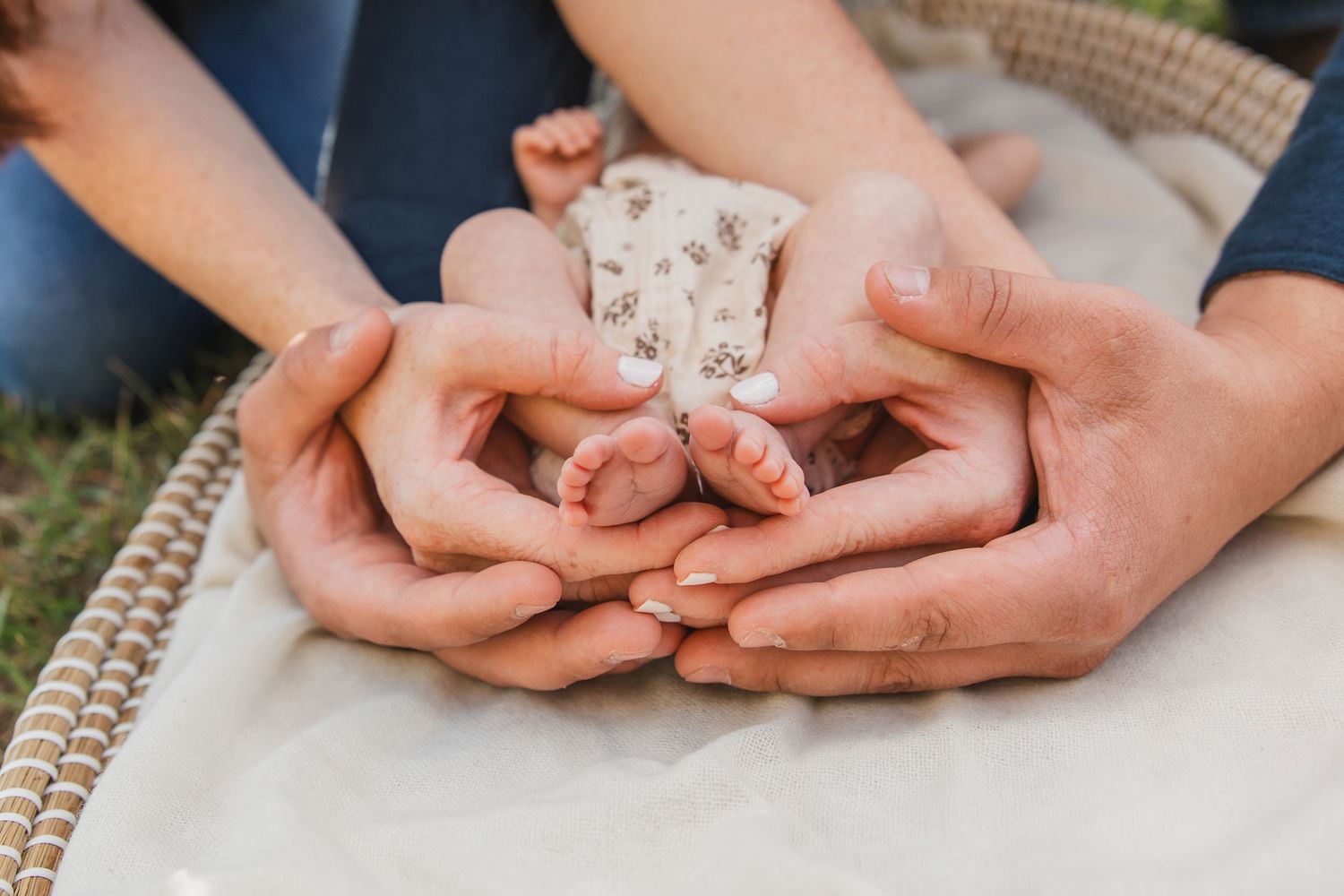 Parents cradling their newborn baby's tiny feet in their hands outdoors on a soft blanket.