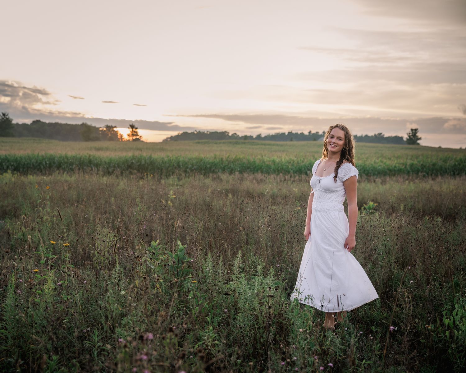 A young woman in a white dress stands smiling in a lush green field at golden hour sunset.