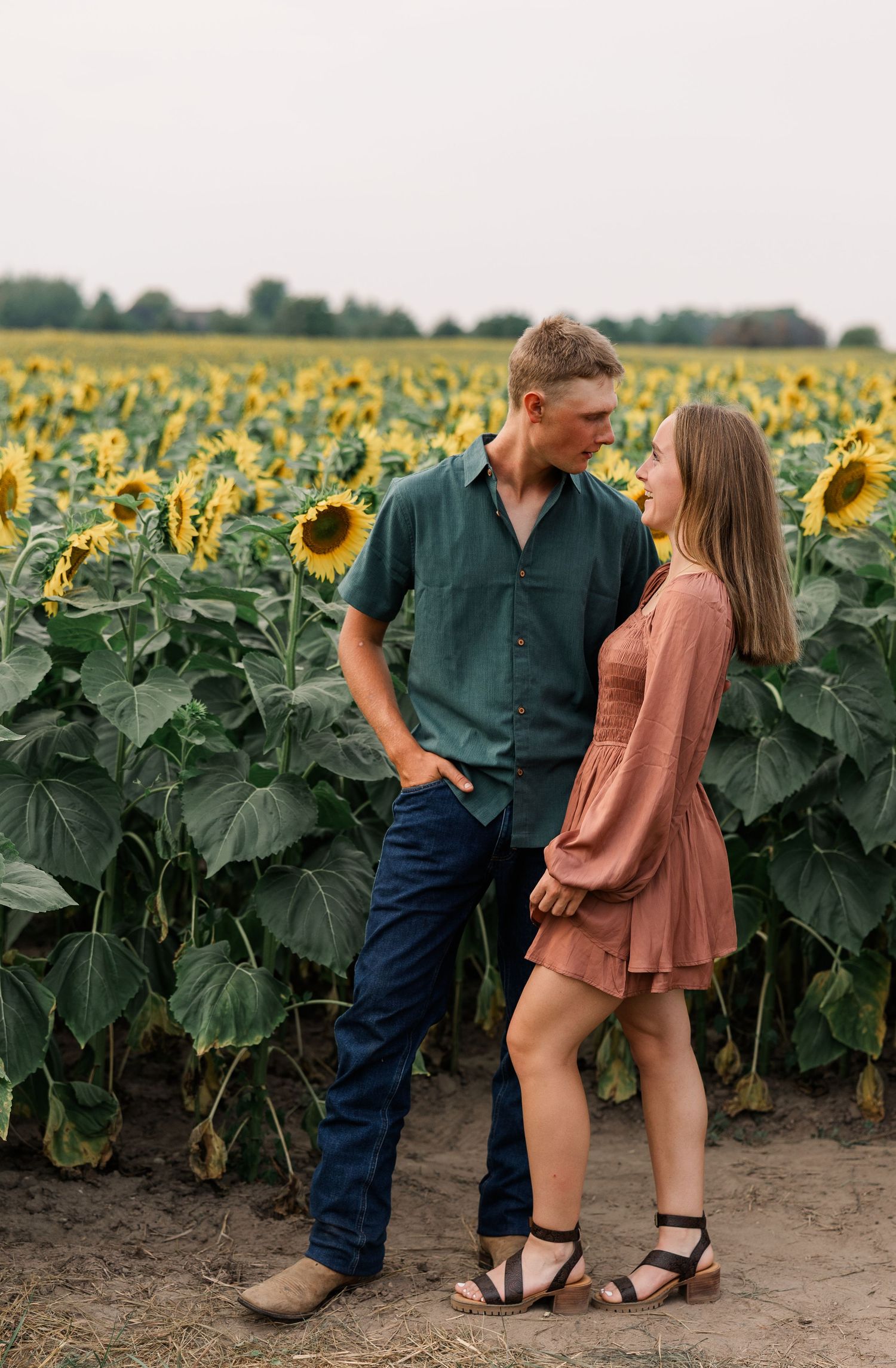 Couple holding hands in a sunflower field, man in green shirt and jeans, woman in pink dress and sandals.