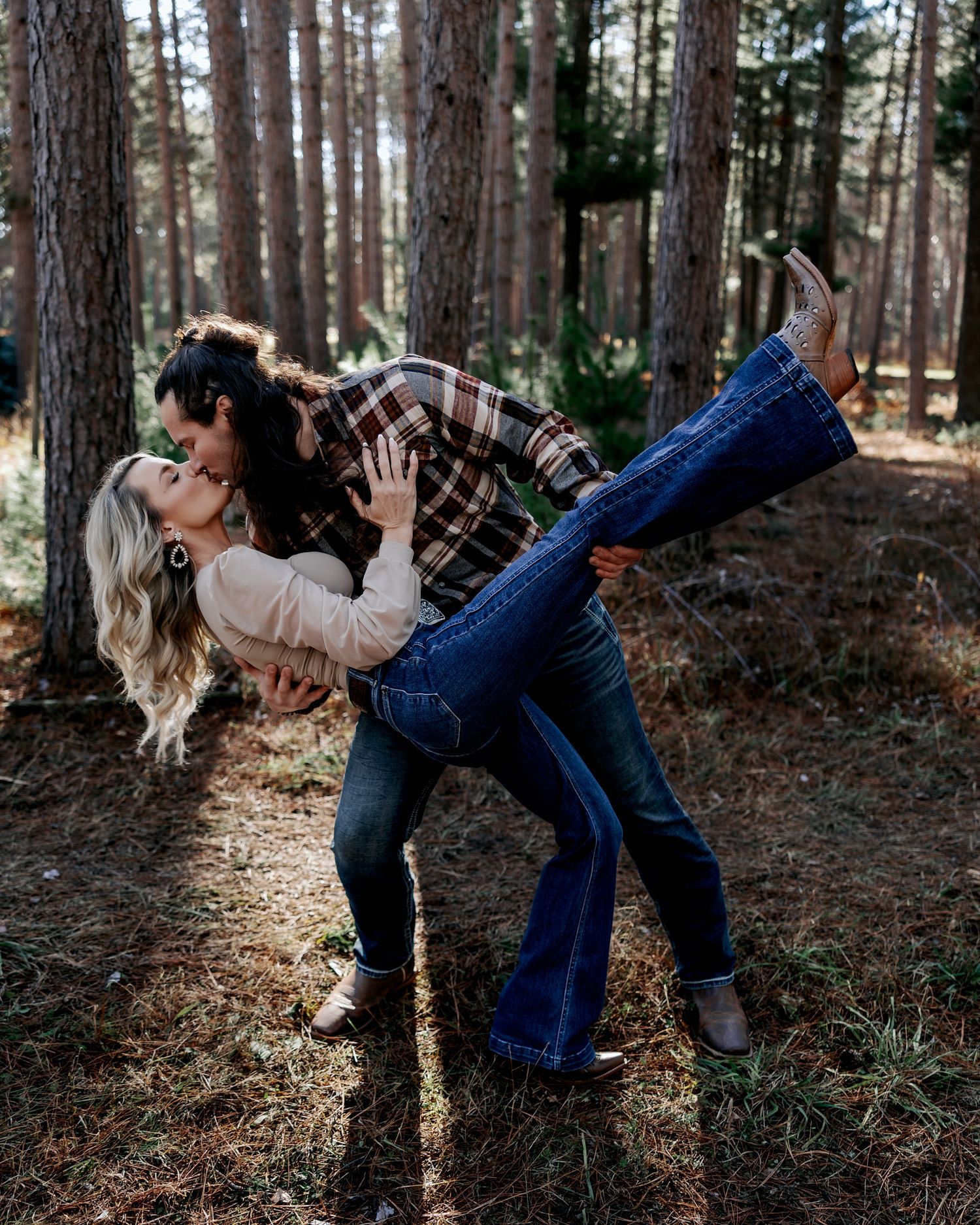 Couple sharing a romantic dip kiss in a pine forest, wearing flannel and flare jeans.