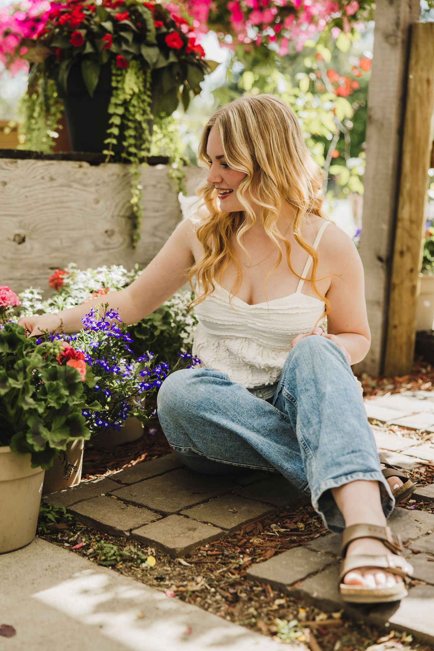 Young woman with blonde hair sitting among colorful flowers in a garden, wearing jeans and a white top.