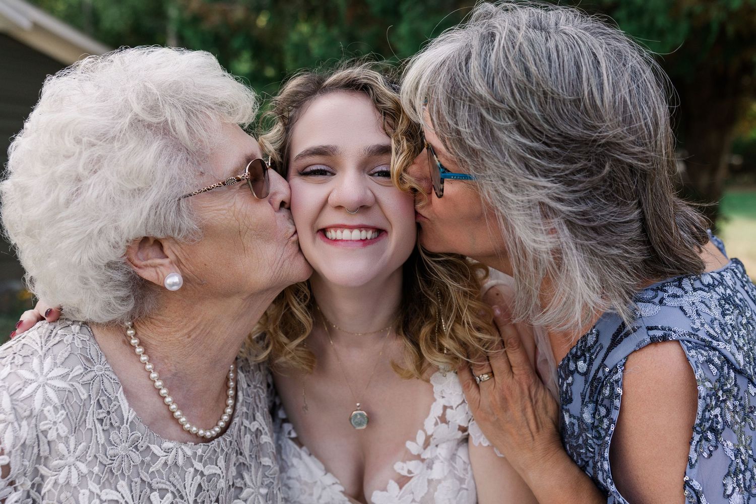 A smiling bride is kissed on the cheeks by two older women at a wedding.