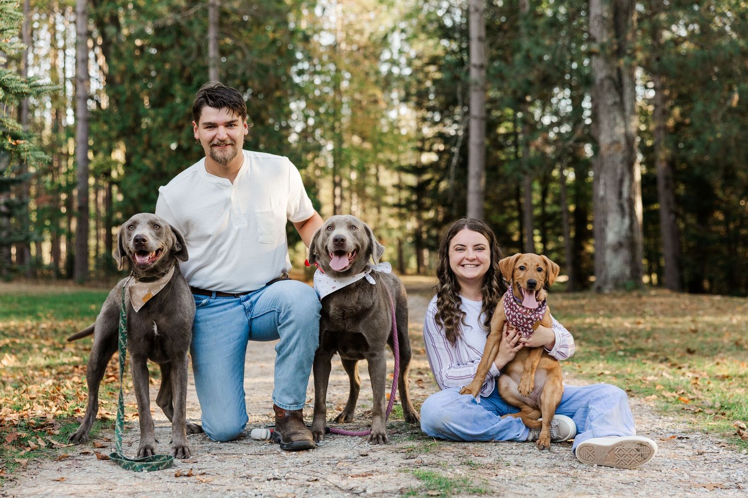 Couple posing outdoors with three dogs, including Weimaraners and a Dachshund, in a wooded park setting.