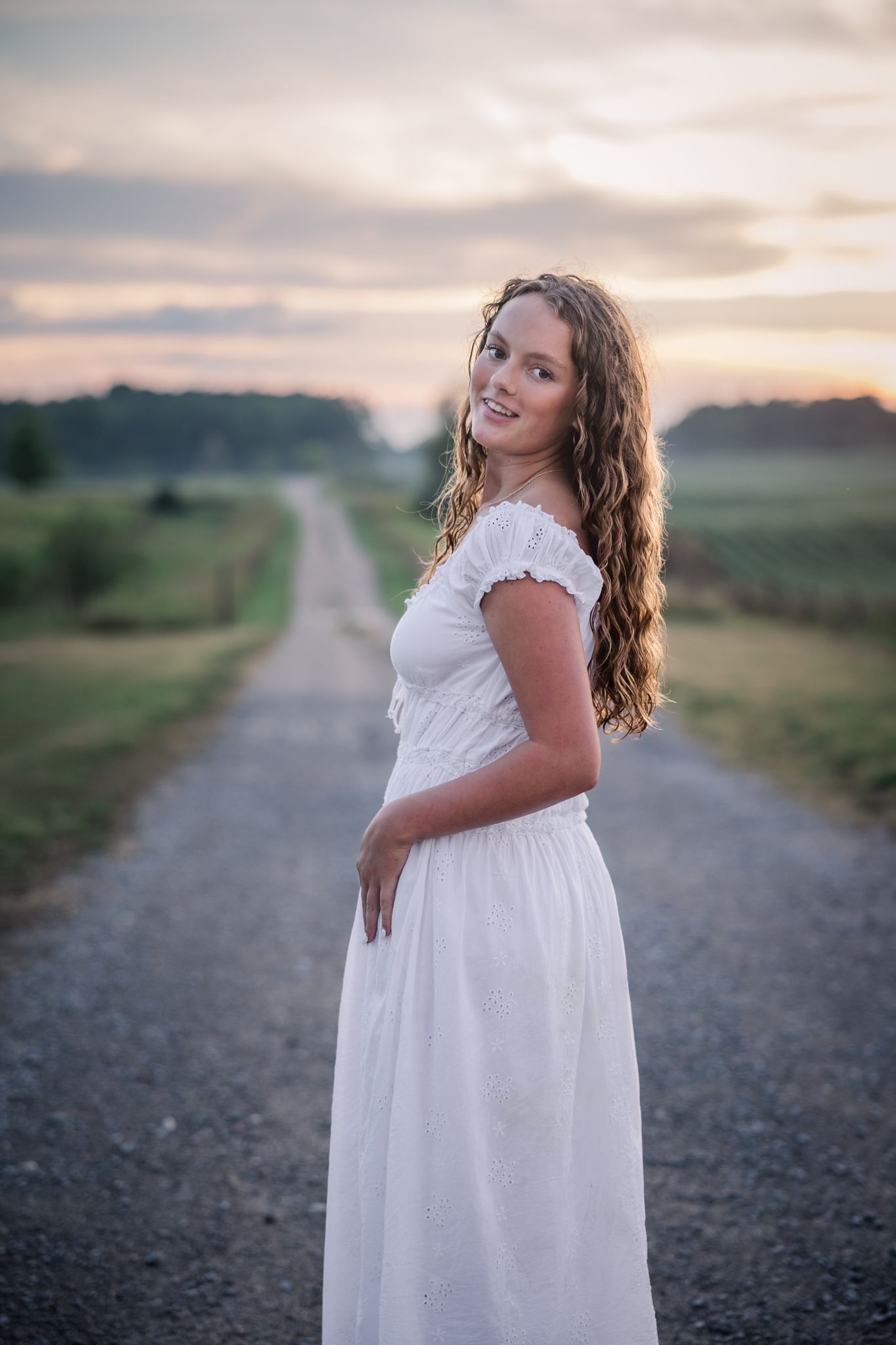 Young woman in white off-shoulder dress standing on rural dirt road at sunset with curly hair and warm smile.
