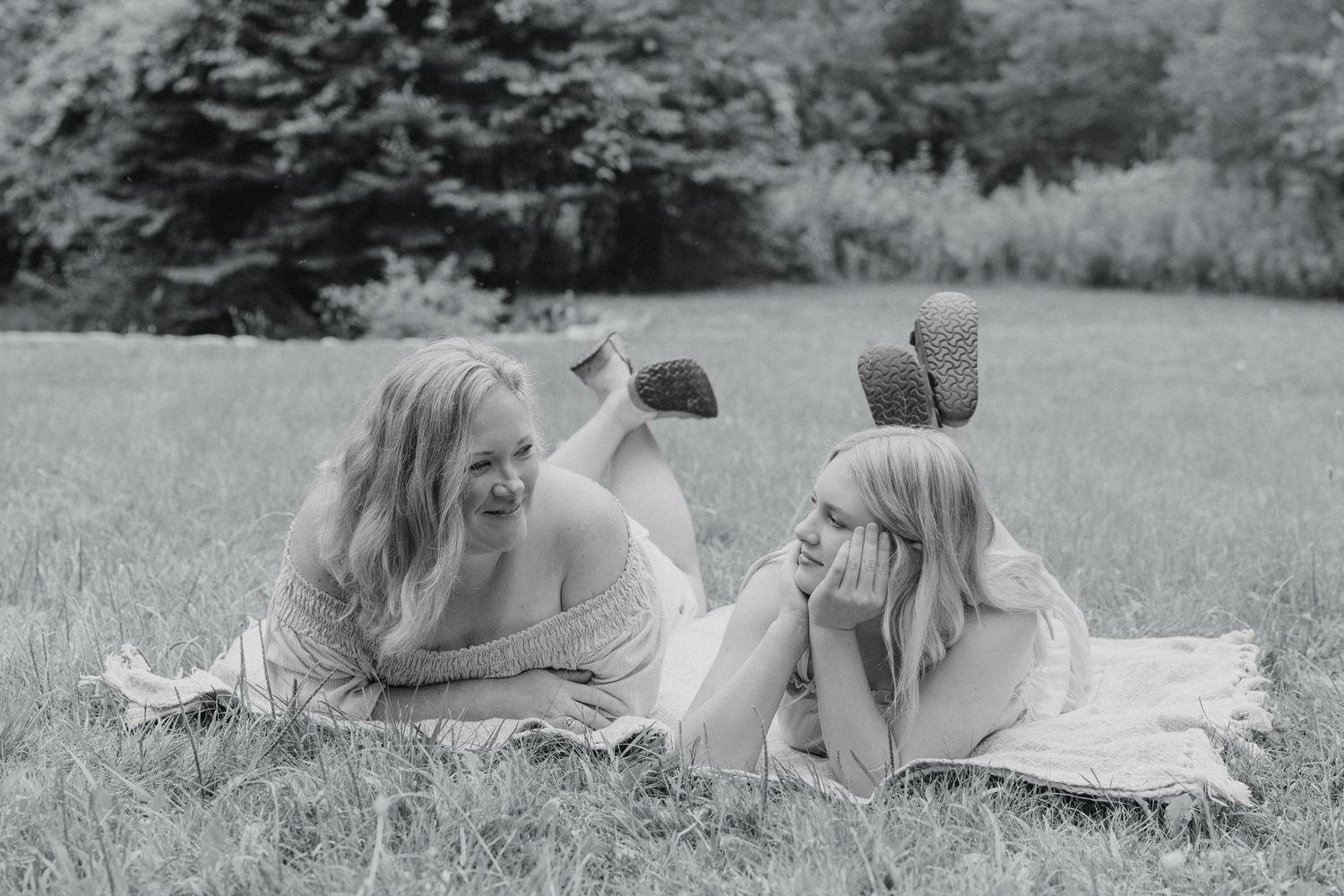 Three women lying on a blanket in a grassy field, laughing together in a black and white outdoor photo.