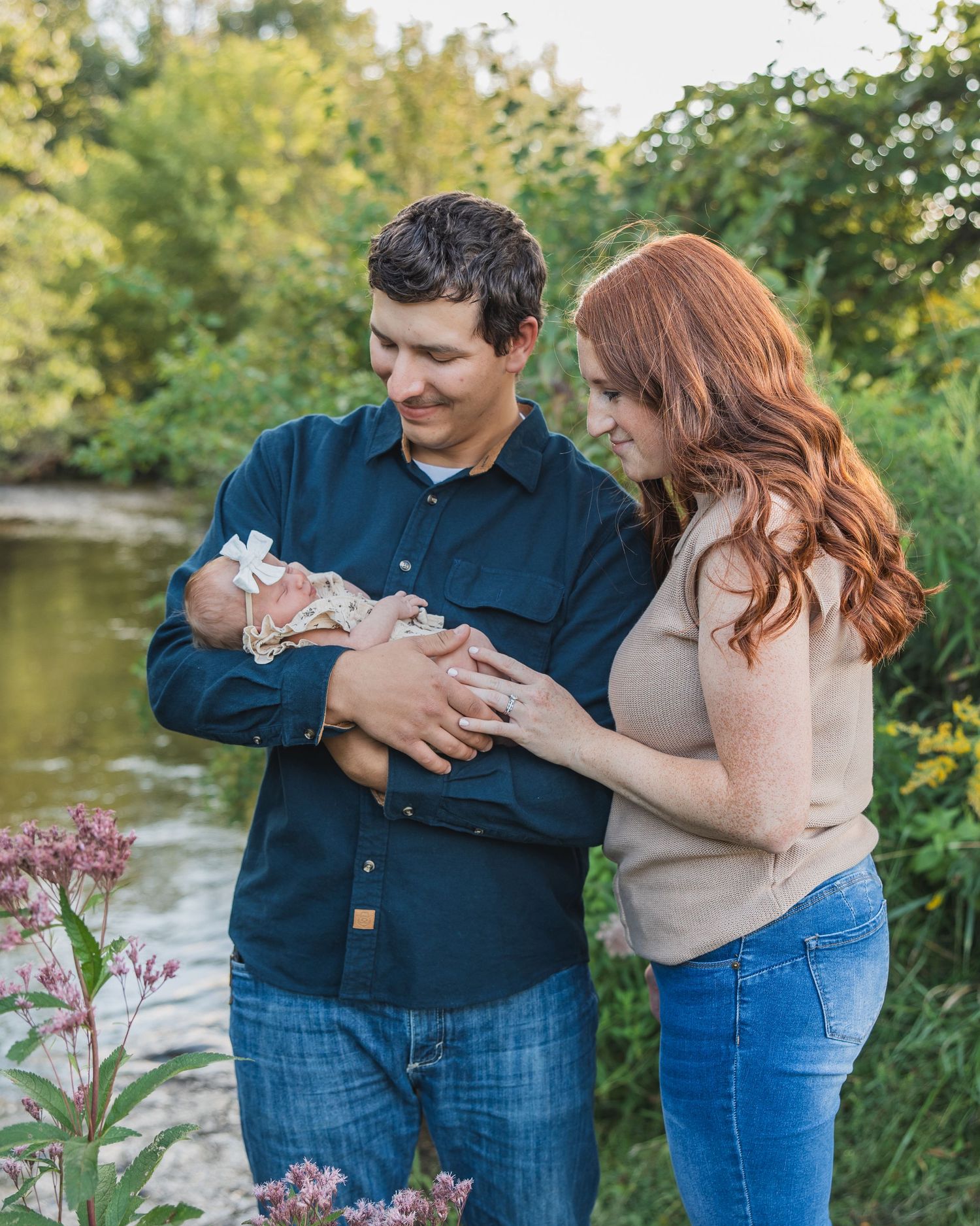 Young couple smiling at newborn baby outdoors near a creek surrounded by lush green trees and pink wildflowers.