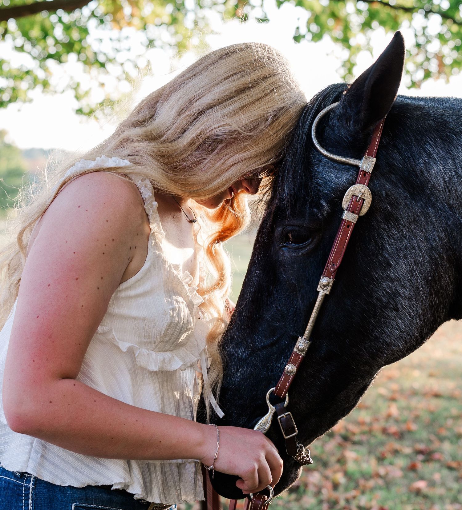 Blonde girl leans forehead against black horse, sharing a tender moment outdoors in soft light.
