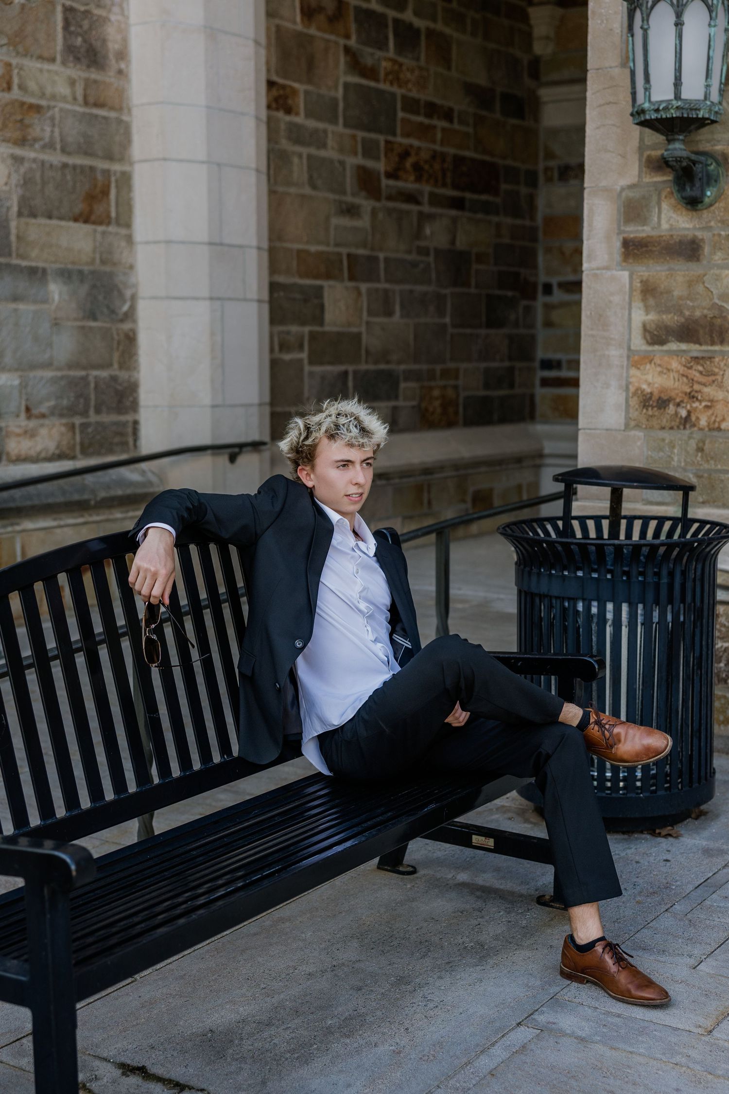 Young man in dark suit and brown loafers sitting relaxed on a black bench against a stone building exterior.