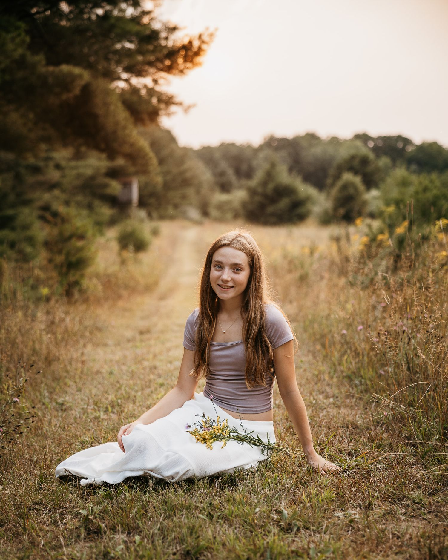 Young woman sitting in a grassy field path holding wildflowers, wearing white skirt and gray top at golden hour.