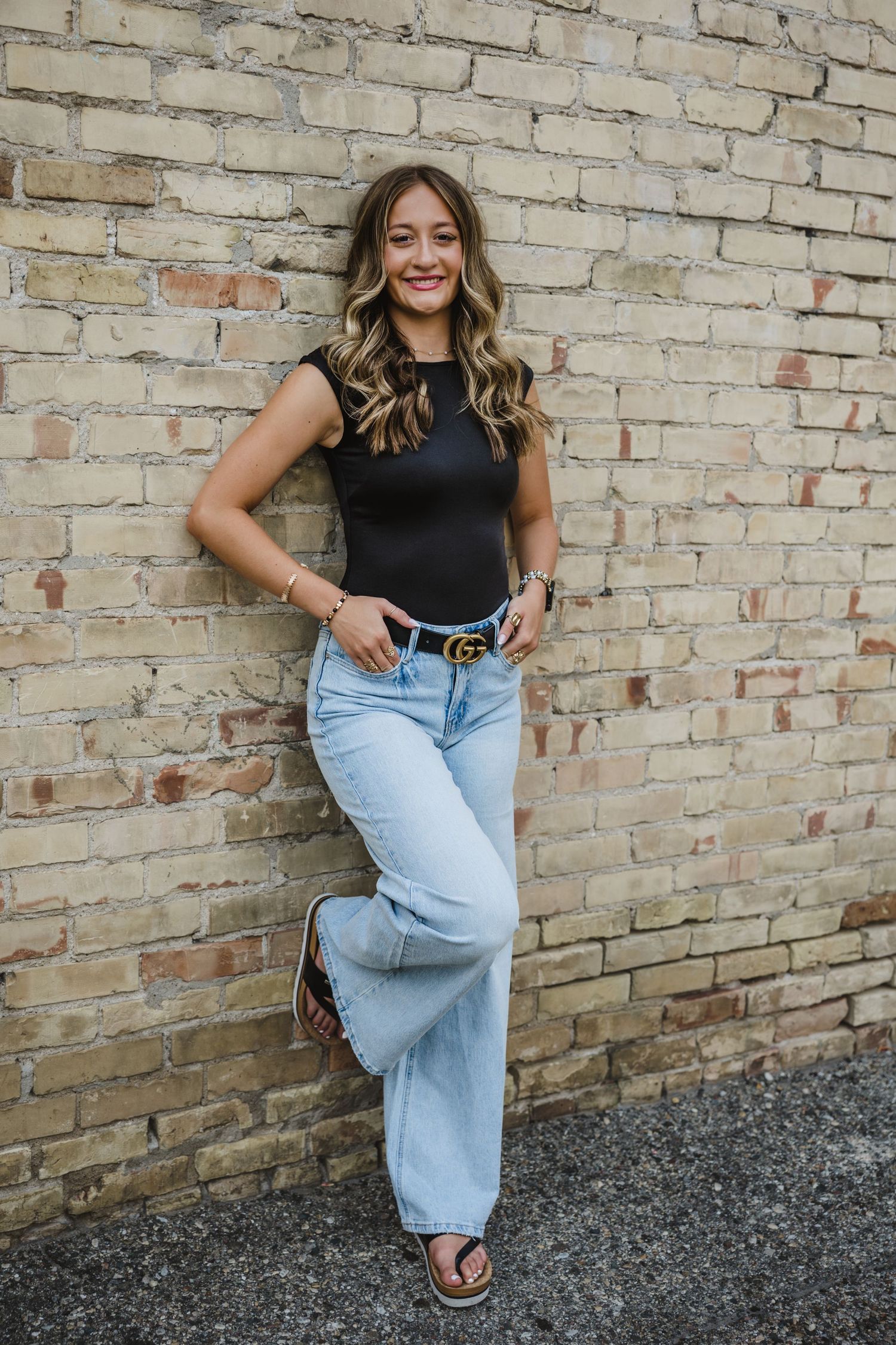 Young woman in black top, light wash wide-leg jeans, and Gucci belt leaning against a brick wall.